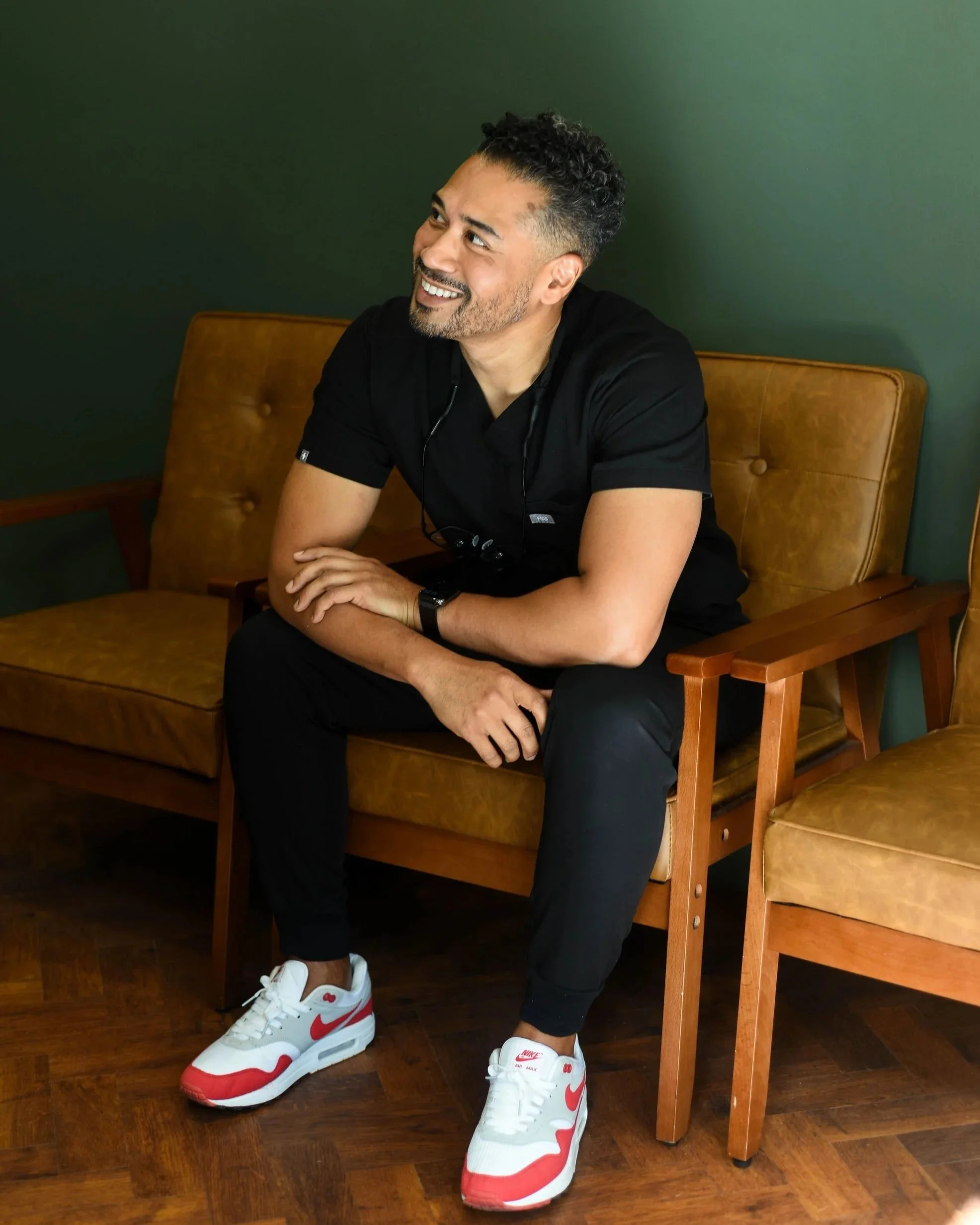 A man sitting on a mustard-colored chair with wooden arms against a dark green wall, smiling and looking to the side, wearing black scrubs and white and red Nike sneakers.