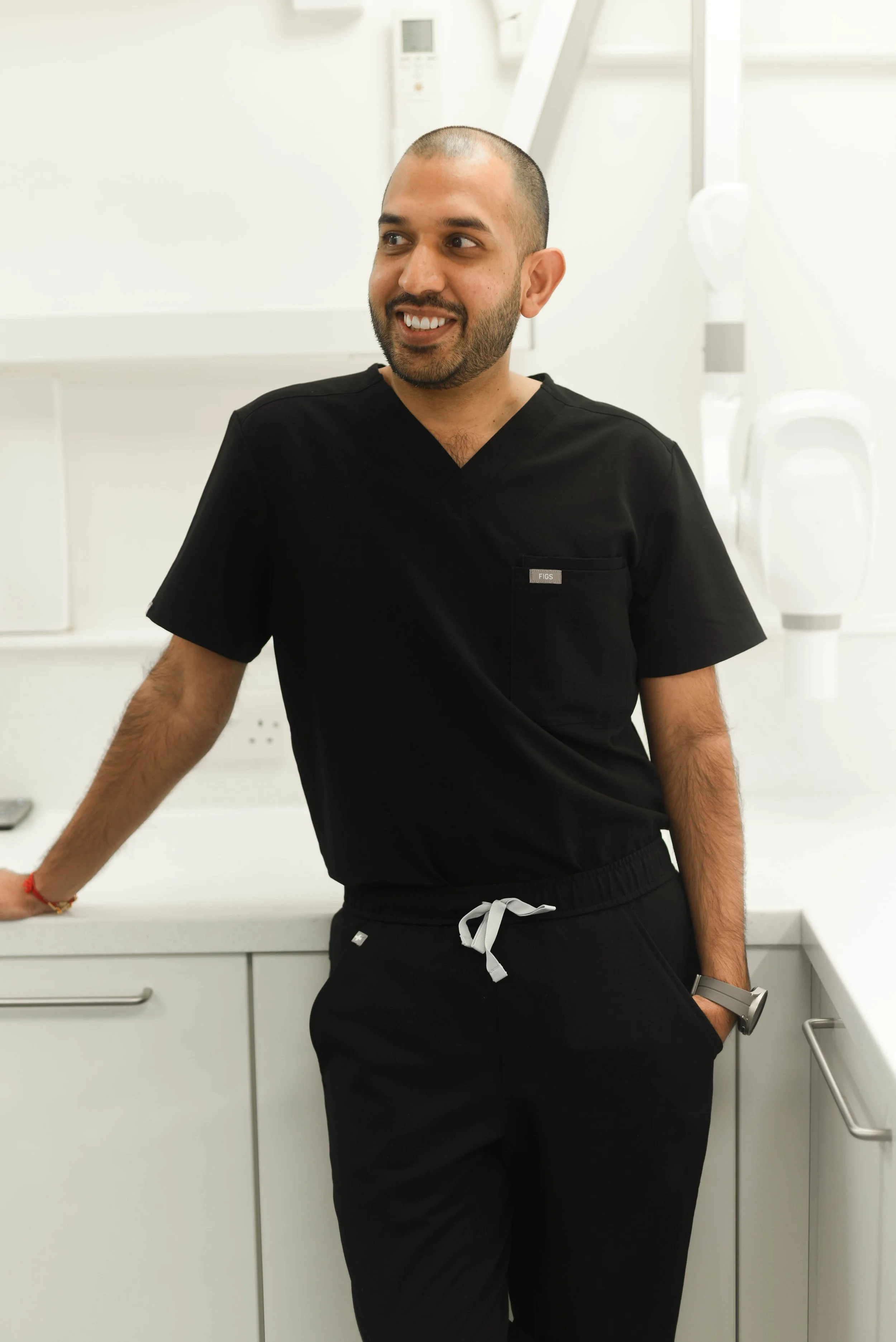 A man with a shaved head and beard wearing black medical scrubs, smiling and leaning against a white countertop in a medical or clinical setting.