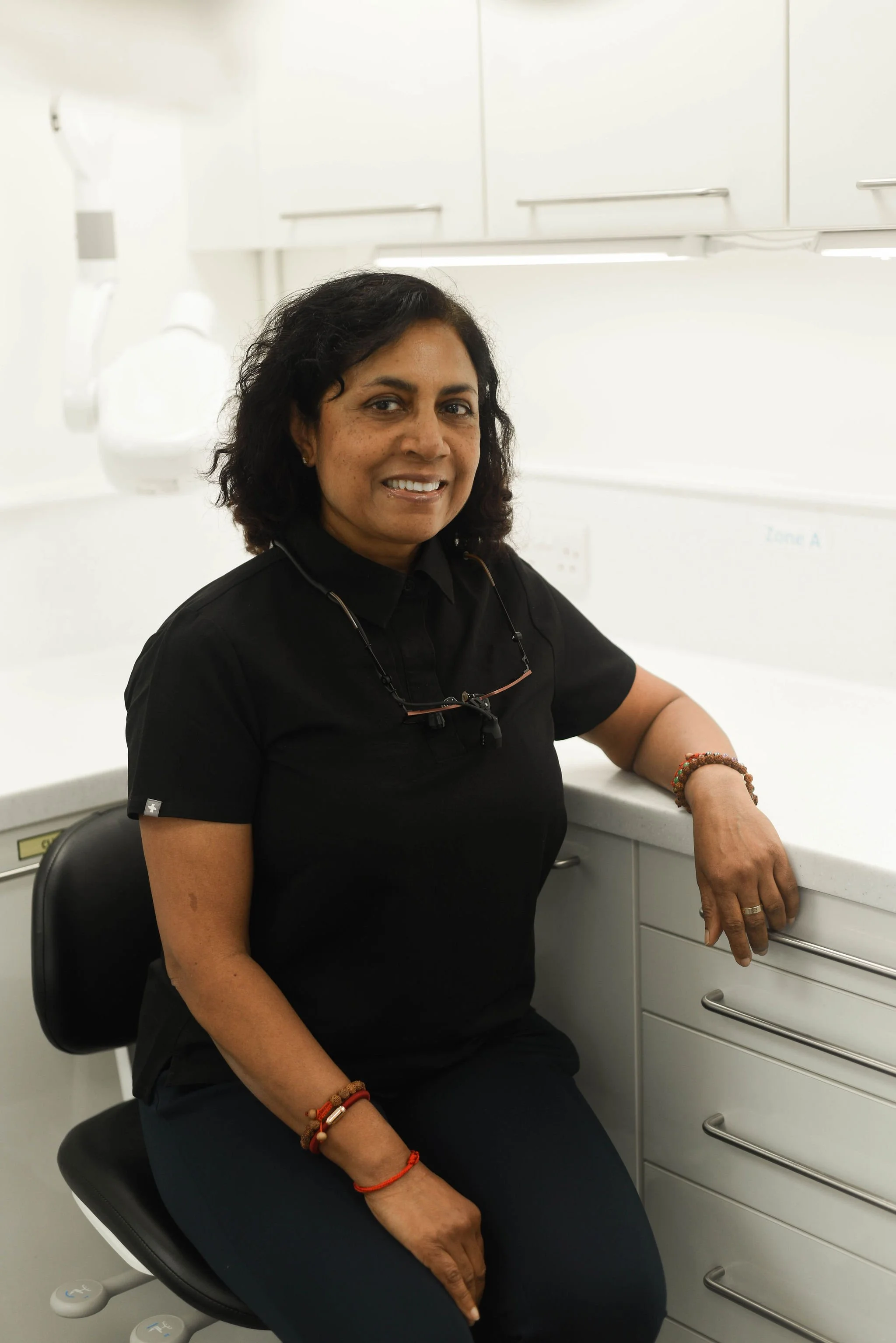 A woman with dark curly hair, wearing a black shirt, sitting in a medical or laboratory office with white cabinets, smiling at the camera.