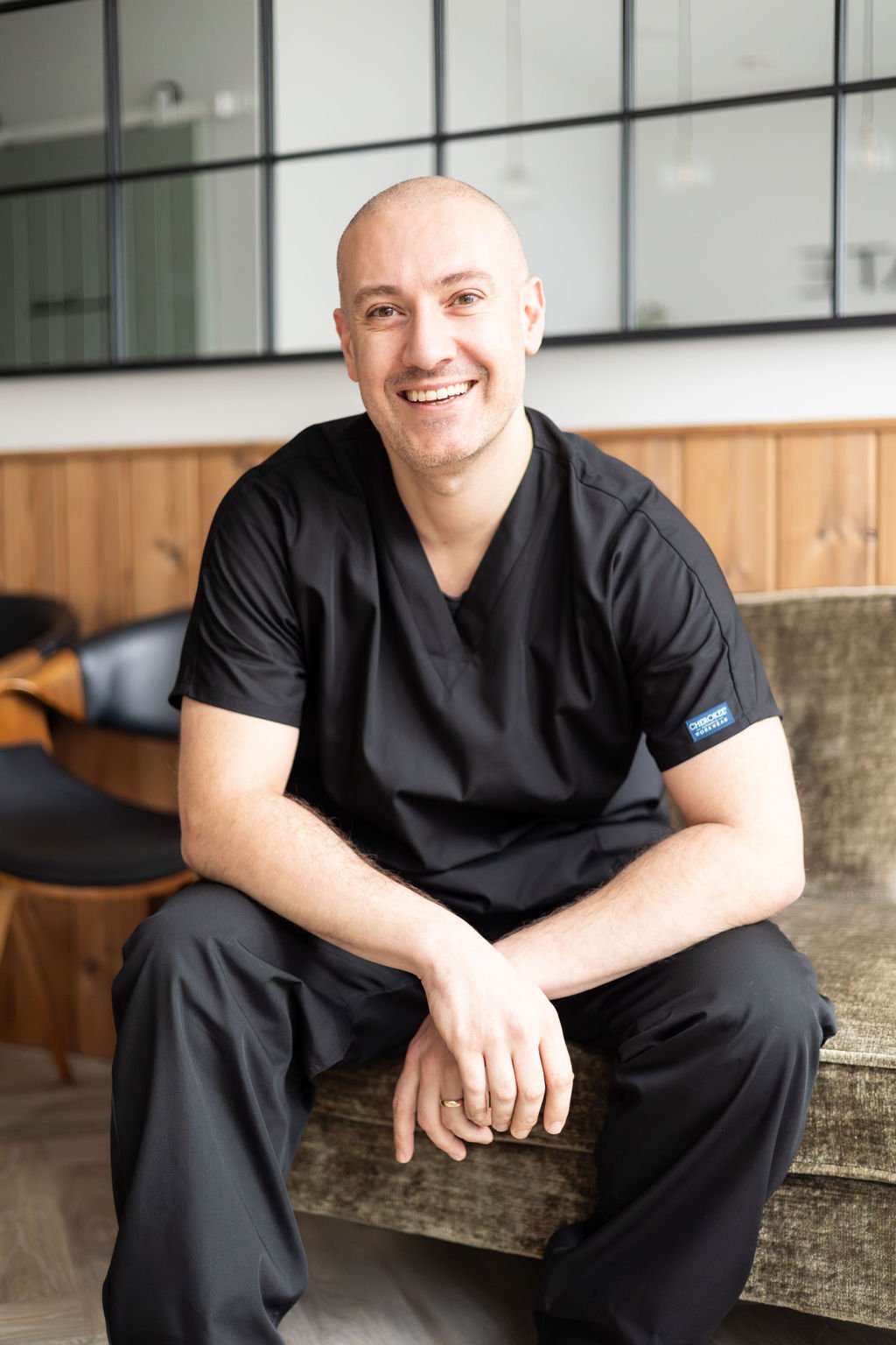 A smiling man in black scrubs sitting on a couch in a modern waiting area with glass windows and wood paneling.