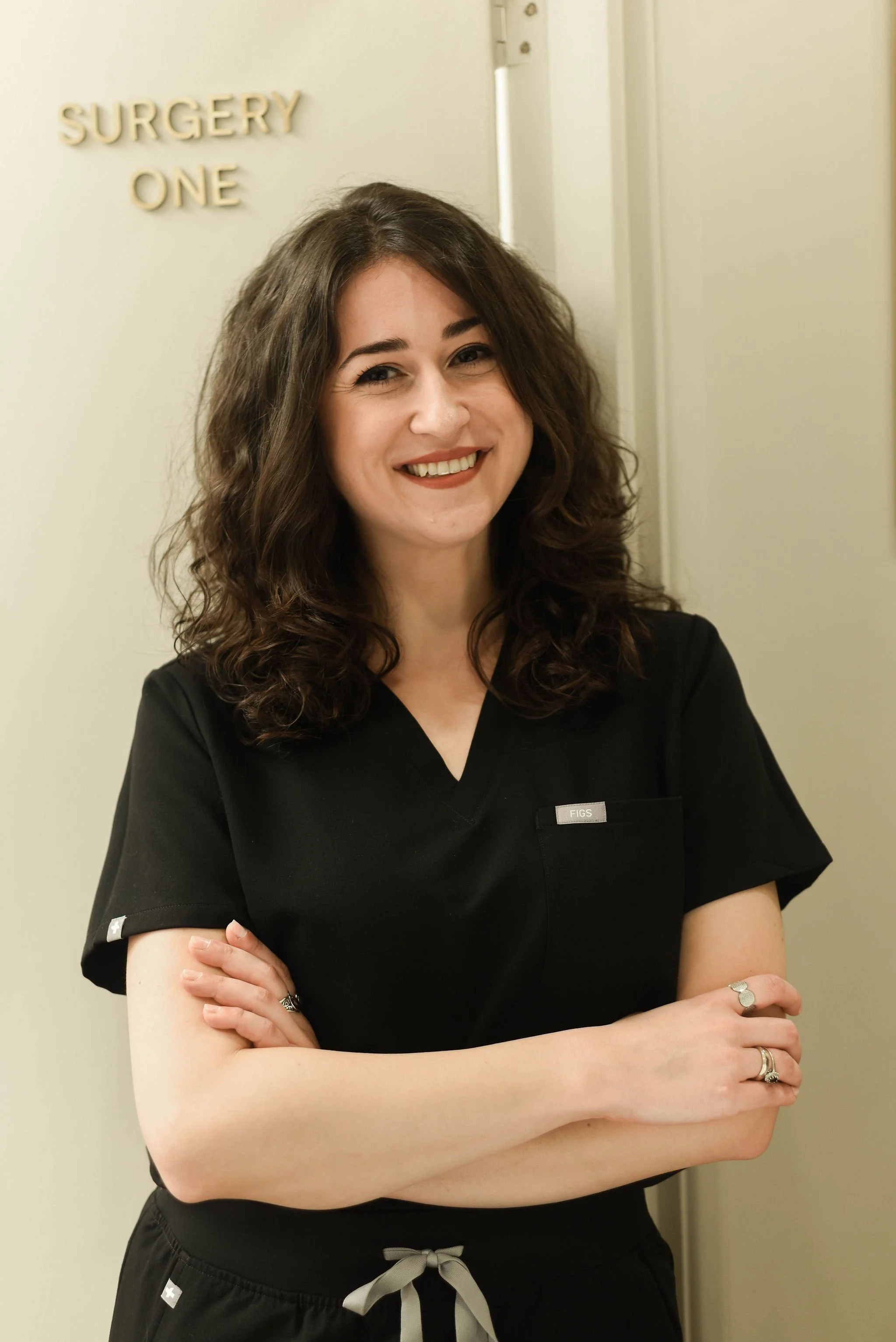 A young woman with curly brown hair smiling, wearing black scrubs, standing in a room with a sign that says 'SURGERY ONE' on the wall behind her.
