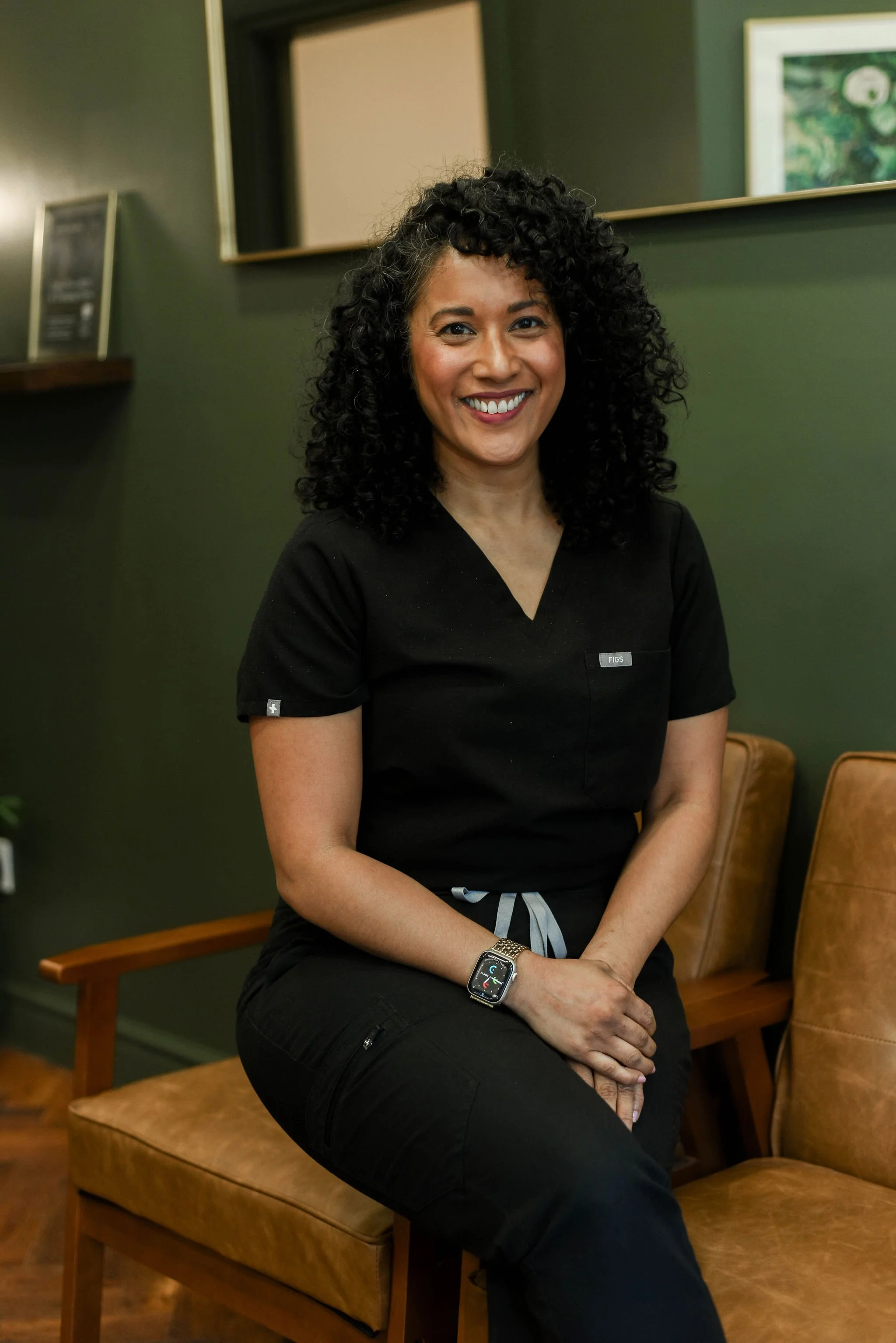 A woman with curly black hair, wearing a black medical uniform, sitting on a tan chair in a room with dark green walls and framed art, smiling at the camera.