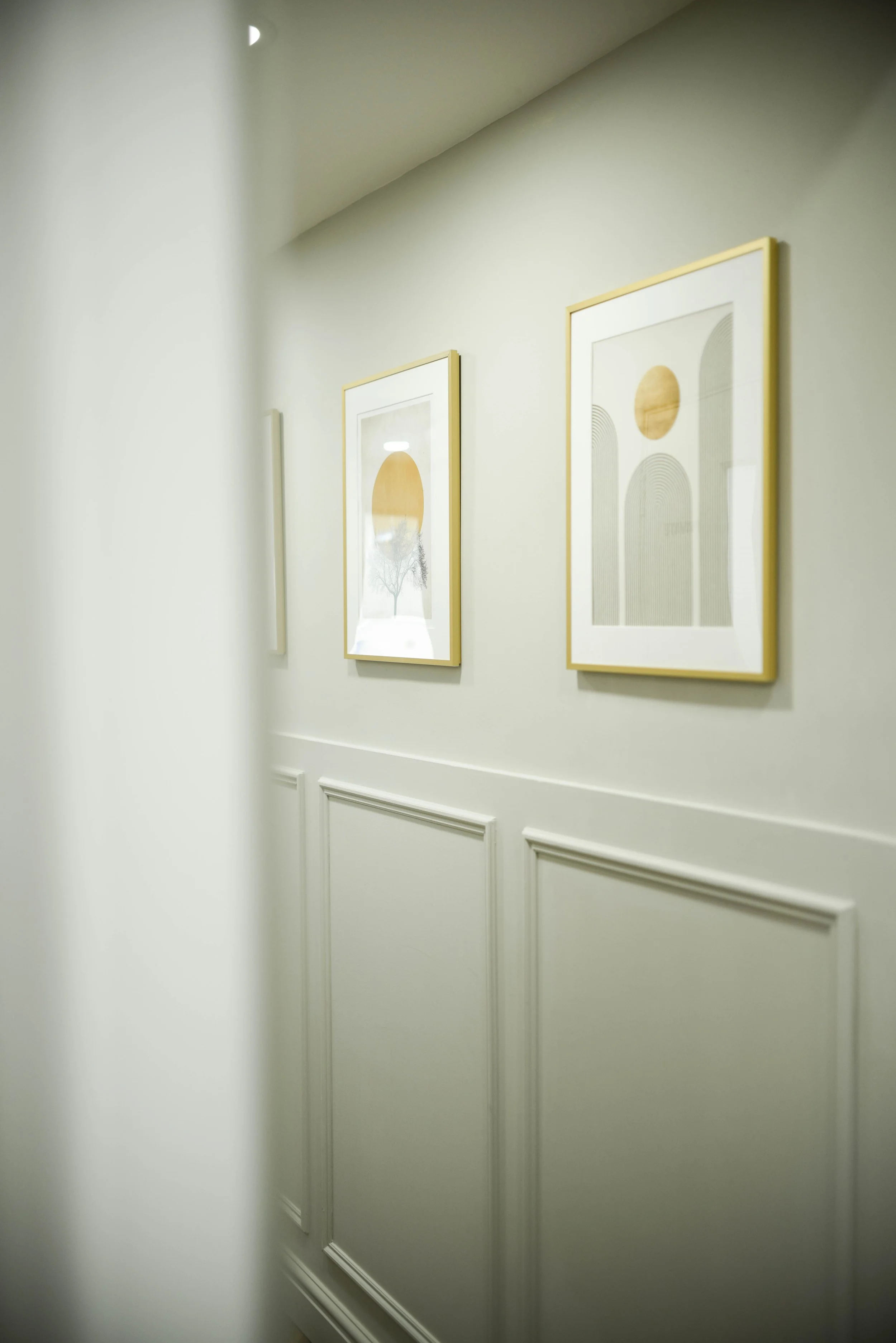 Part of an art gallery wall with three framed abstract prints featuring circles and arches, with a white wainscot wall below, seen through a slightly blurred foreground on the left.