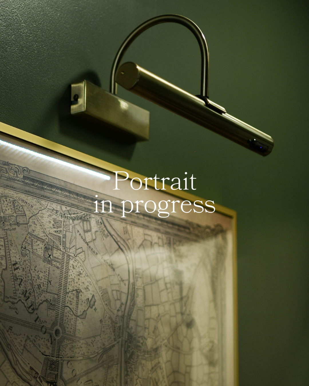 A vintage picture light illuminating a historical map on a green wall, with overlay text 'Portrait in progress'.