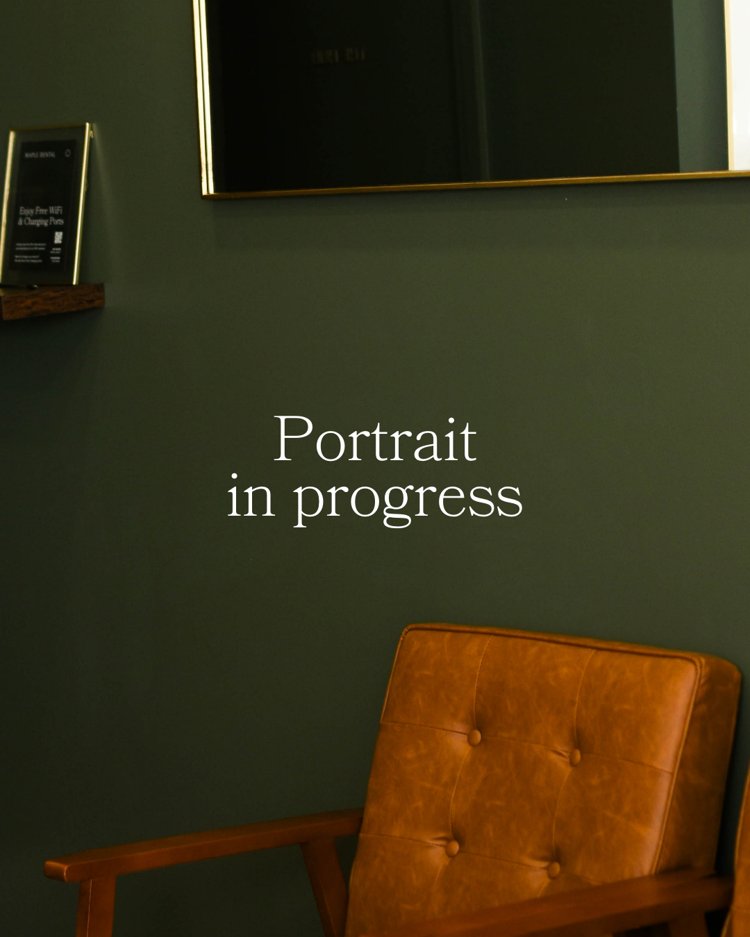 A waiting area with a brown leather chair against a dark green wall, a small shelf with a black frame sign, and a large mirror reflecting a door.