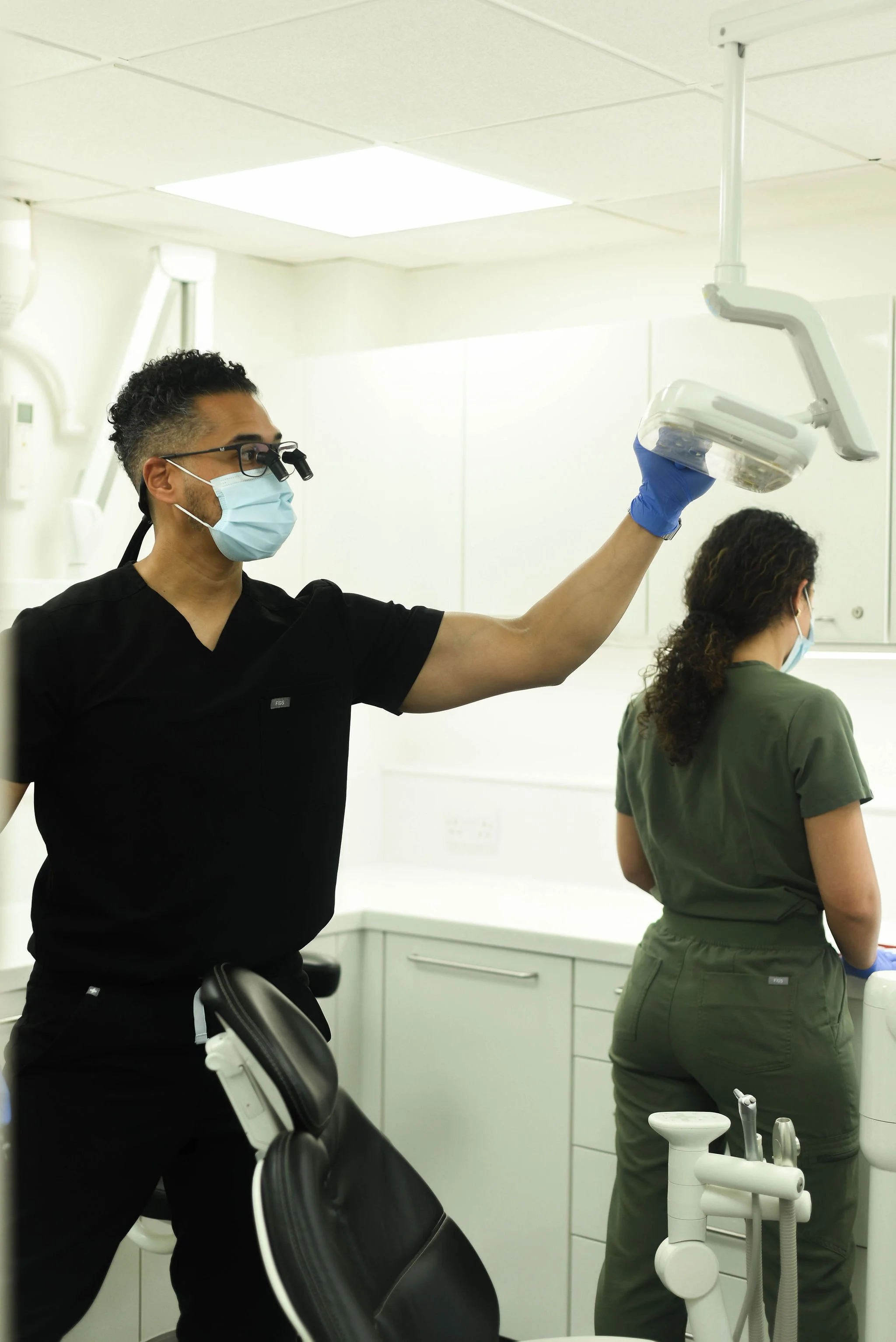 A male dentist wearing a face mask, magnifying glasses, and blue gloves is lifting a light over a dental chair. A female patient wearing a face mask and green scrubs is seated with her back to the camera in a dental office.