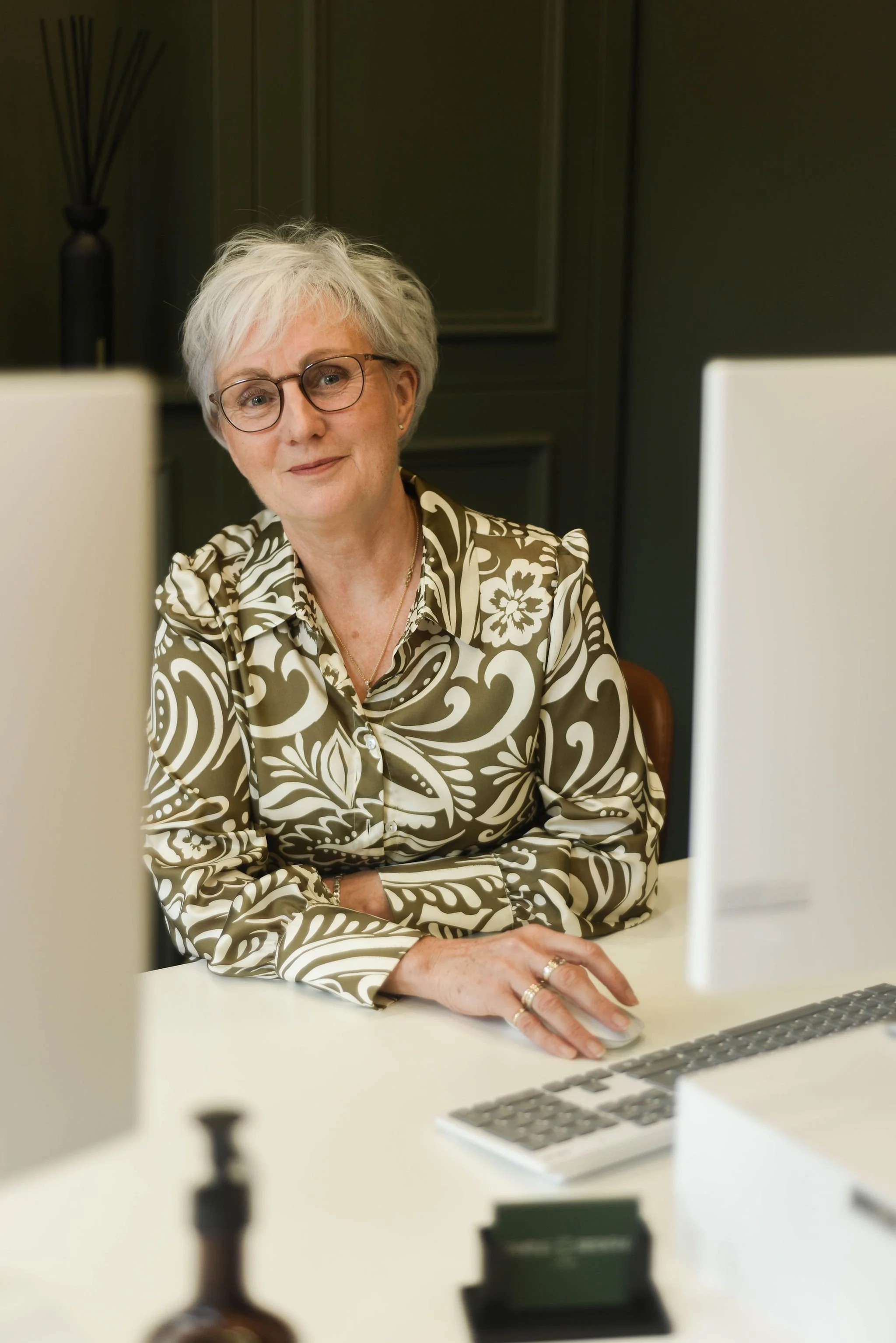 Older woman with glasses and short gray hair working at a desk with two computers, wearing a patterned blouse.