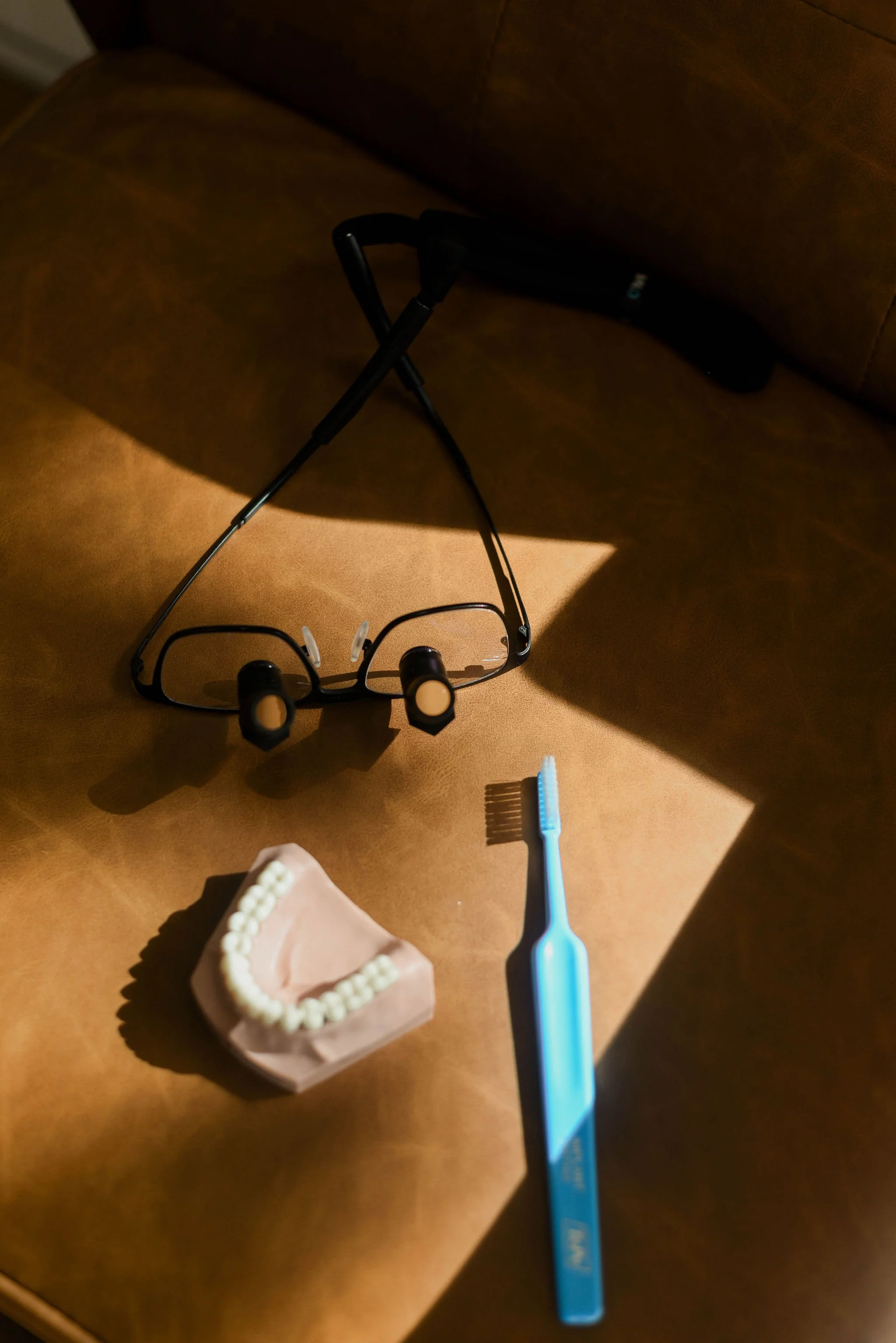 A pair of eyeglasses, a toothbrush, a dental mold with teeth, and a headlamp are arranged on a wooden surface with sunlight casting shadows.