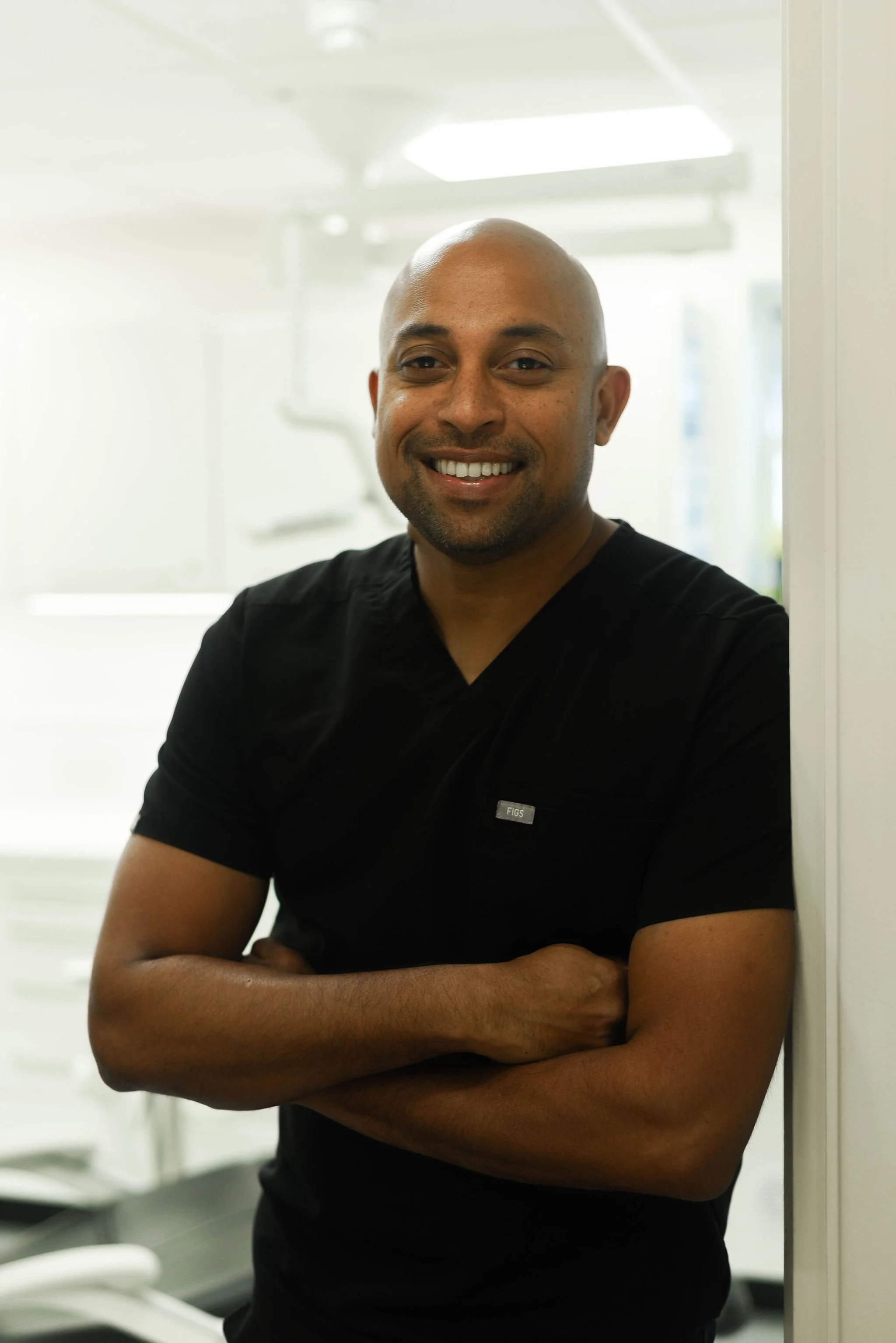 A dentist with a bald head and a goatee, wearing a black scrub top, standing in a modern dental office with blinds and dental equipment in the background, smiling with arms crossed.