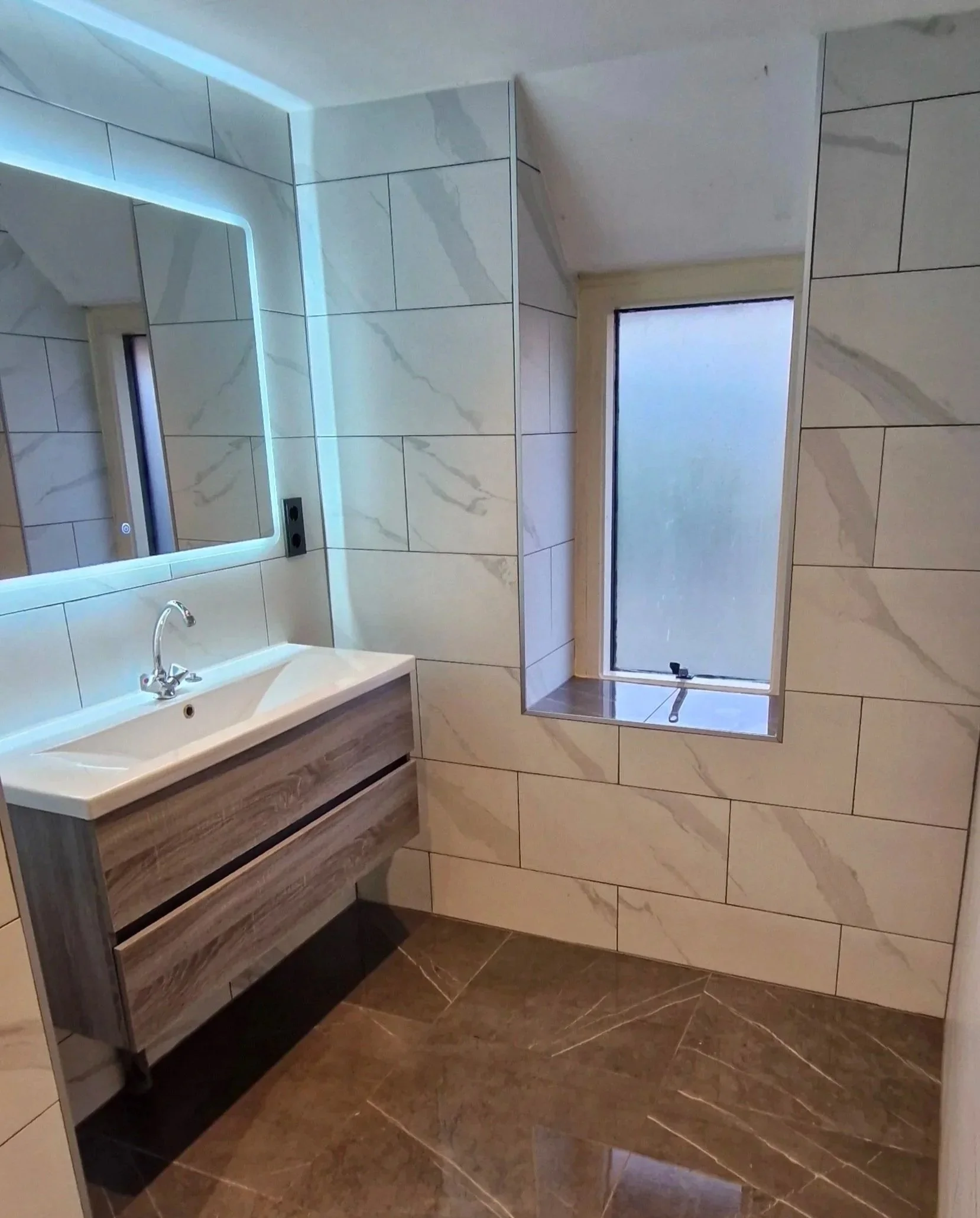Bathroom with large cream marble tiles on walls and brown marble tiles on floor, a rectangular wall-mounted sink with a wood grain finish, a mirror with backlighting above the sink, a window with frosted glass, and a smaller window in a wall niche.