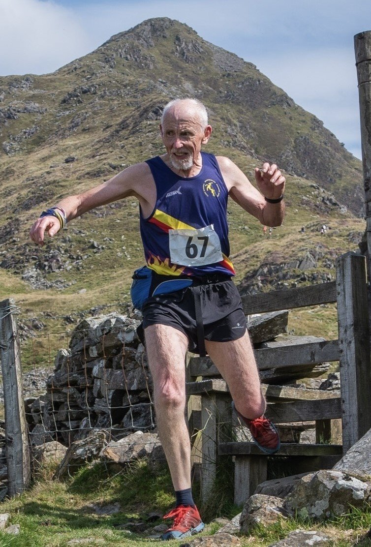 An elderly man running outdoors on a rocky trail with mountains in the background, wearing a race bib number 67, athletic gear, and red running shoes.
