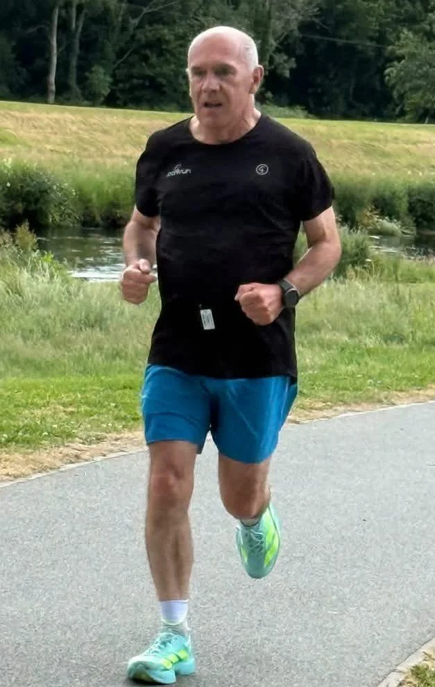 A man running outdoors on a paved path near a grassy area with trees and a small pond in the background.