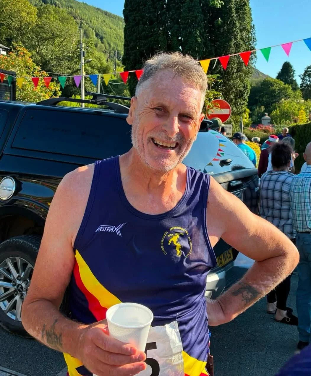An older man with gray hair and a beard smiling, holding a white cup, wearing a blue athletic tank top with red and yellow accents, standing outdoors at a race event with colorful flags and a black vehicle behind him.