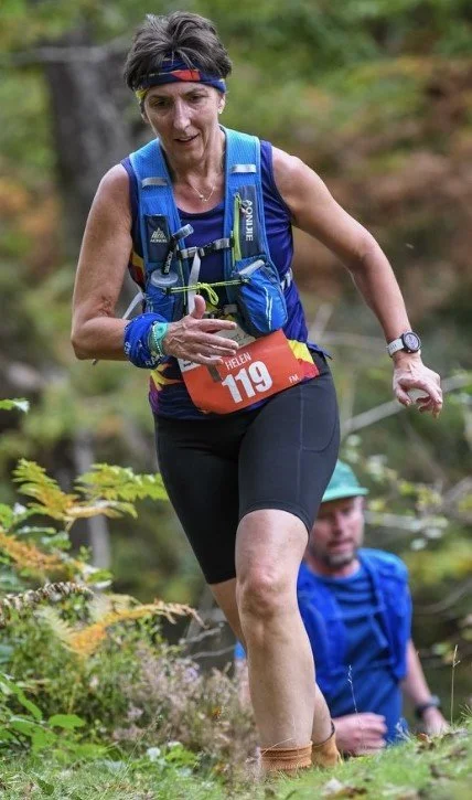 A woman trail running on a forest path, wearing a blue hydration vest, black shorts, and a race bib numbered 119, with another person in a blue shirt in the background.