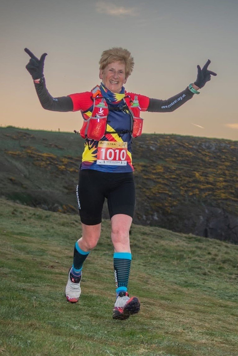 A cheerful woman with short, light hair running outdoors during a race, wearing a colorful athletic outfit, running shoes, and a race bib numbered 1010. She's making a peace sign with both hands, with a scenic landscape in the background.