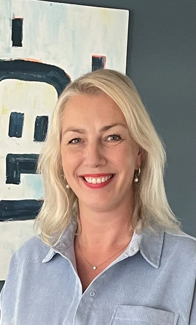 Een lachende vrouw met blond haar en een blauwe blouse die staat voor een schilderij op de muur.