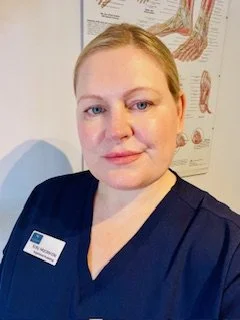 A woman with blonde hair in a bun, wearing a navy blue shirt with a name tag, standing in front of a medical poster showing muscles.