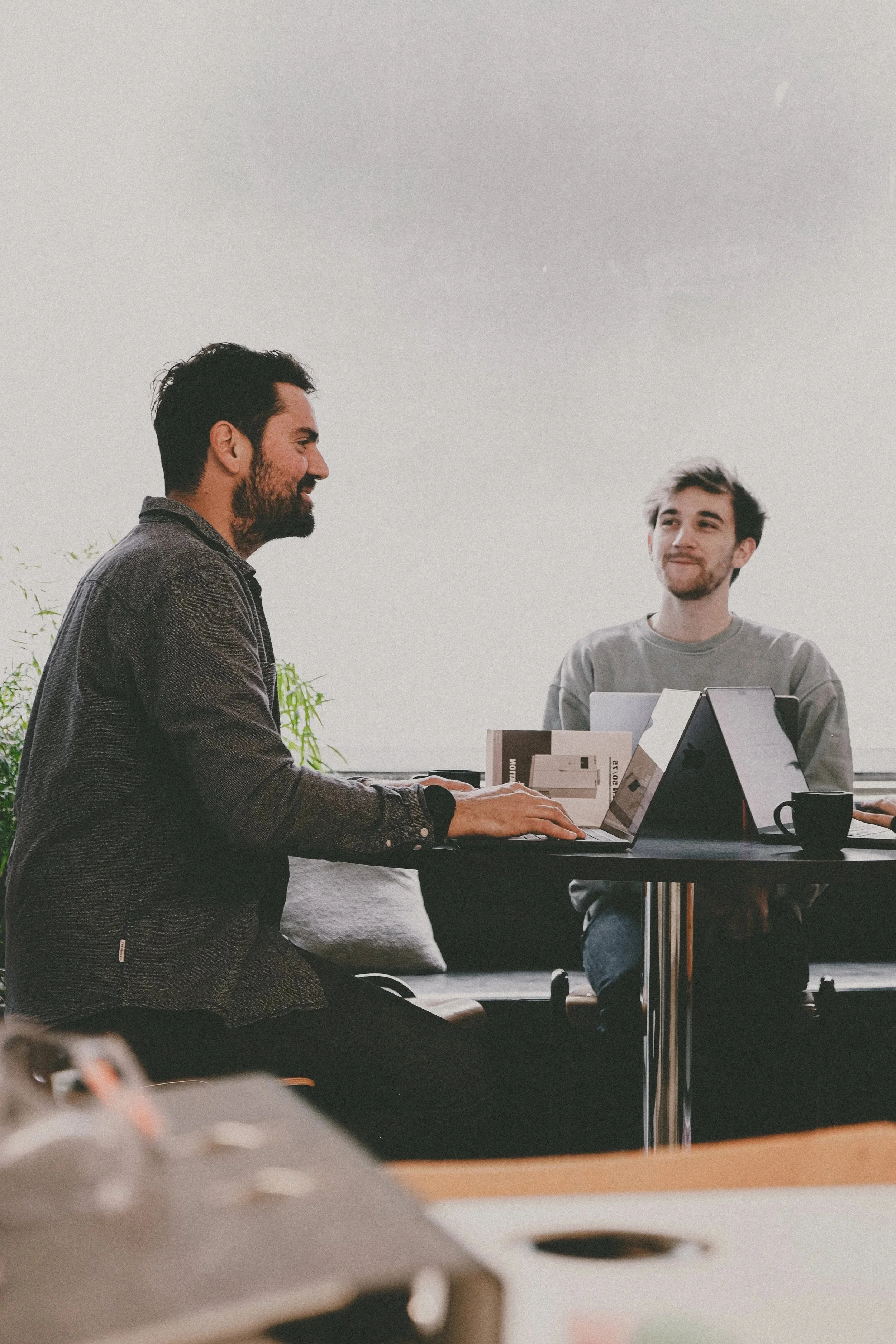 Two men are sitting at a table in a modern office, engaged in conversation. One man has dark hair and a beard, wearing a grey sweater, while the other man has lighter hair, wearing a grey t-shirt. They have laptops and notebooks in front of them.