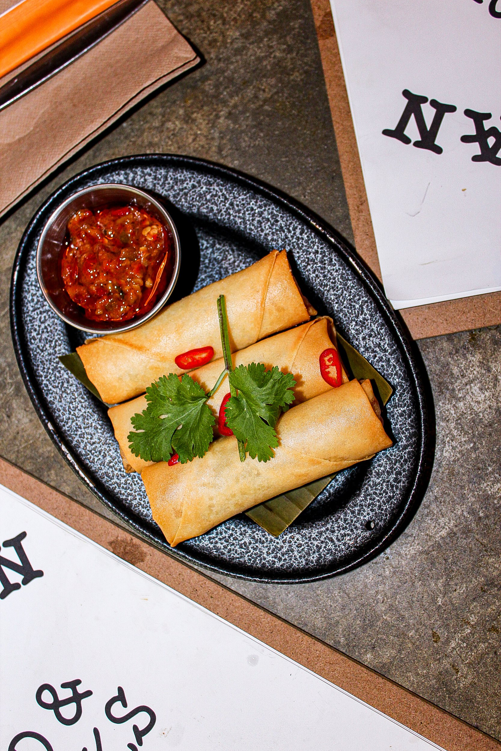 Four spring rolls served on a black oval plate with a side of chili sauce, garnished with cilantro and red chili slices.