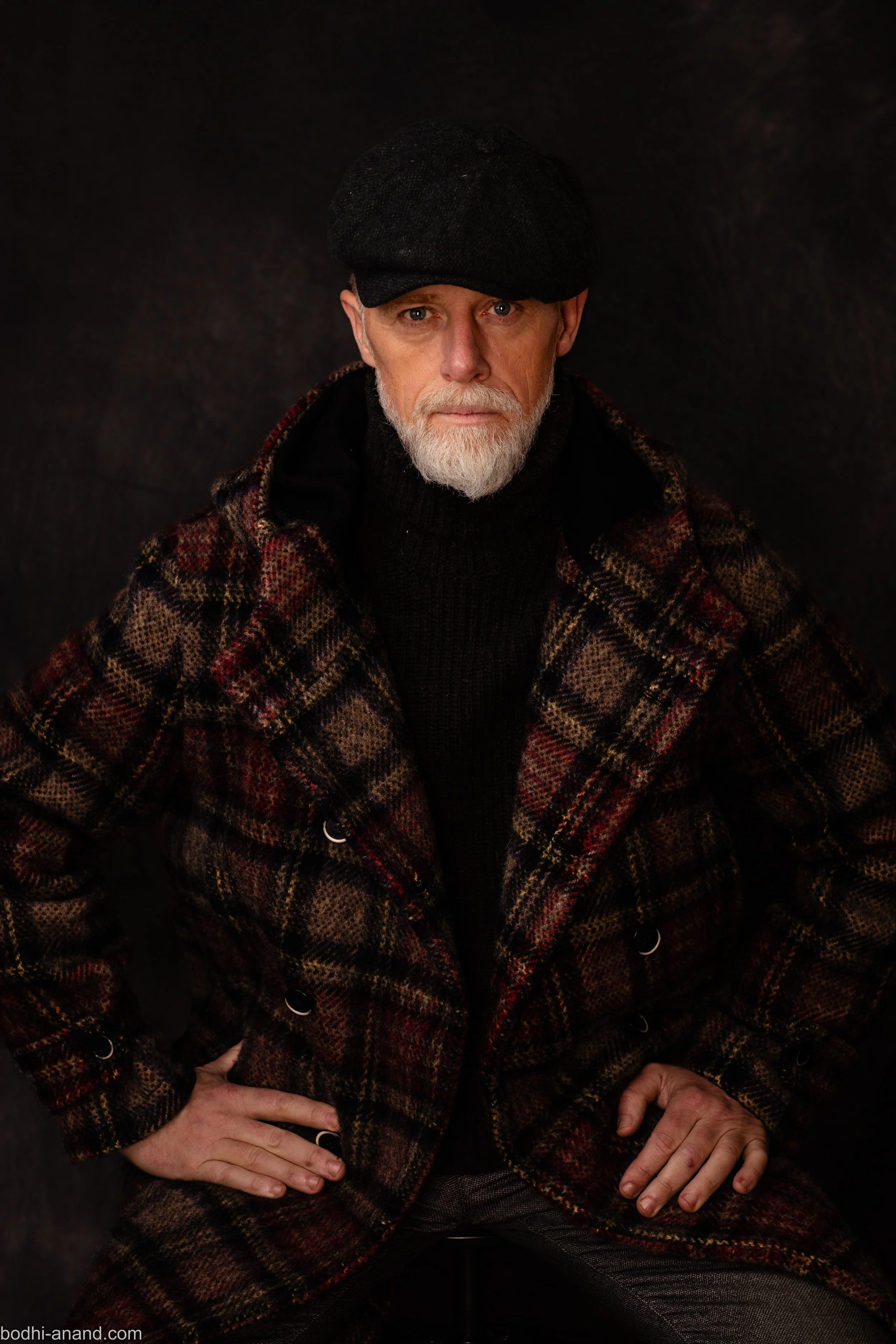 Studio portrait of a white-bearded man wearing a dark cap and checked coat.