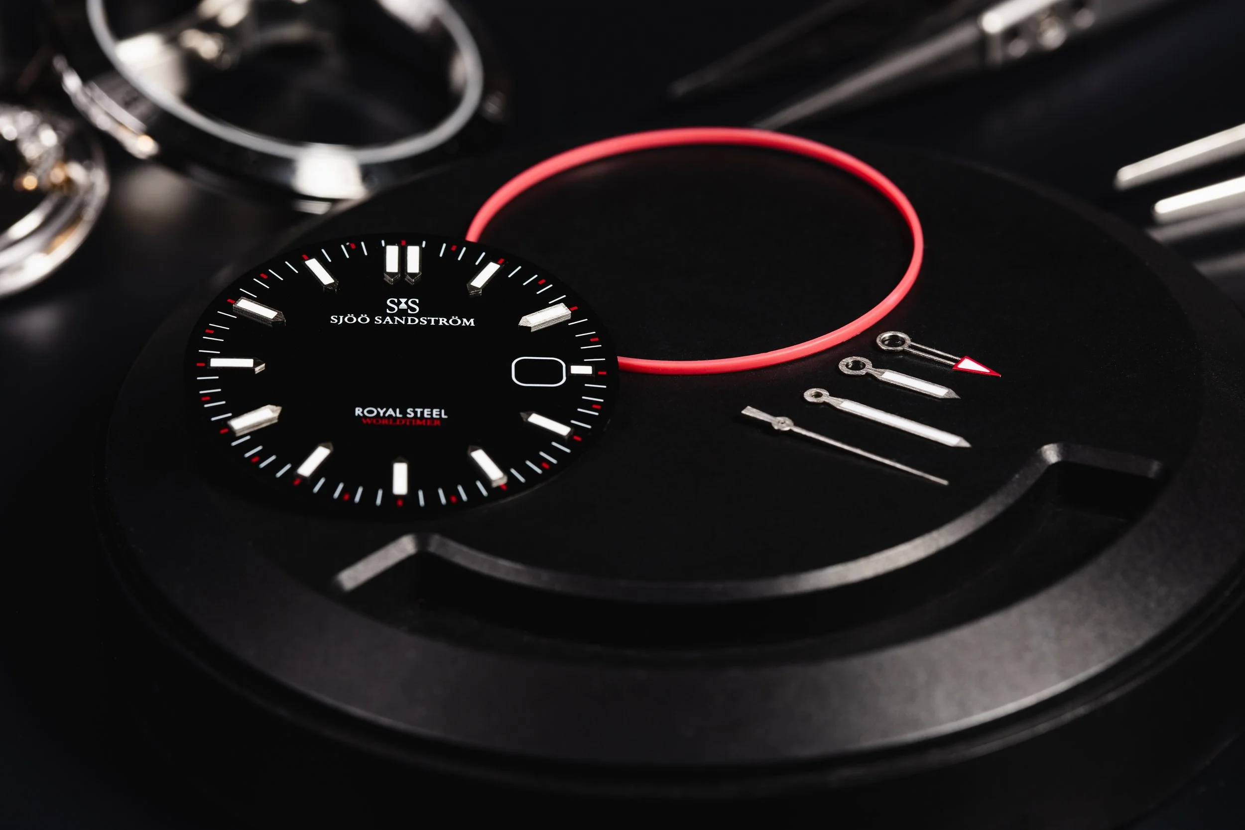 Black Sjöö Sandström watch dial, hands and components arranged on a dark work surface.