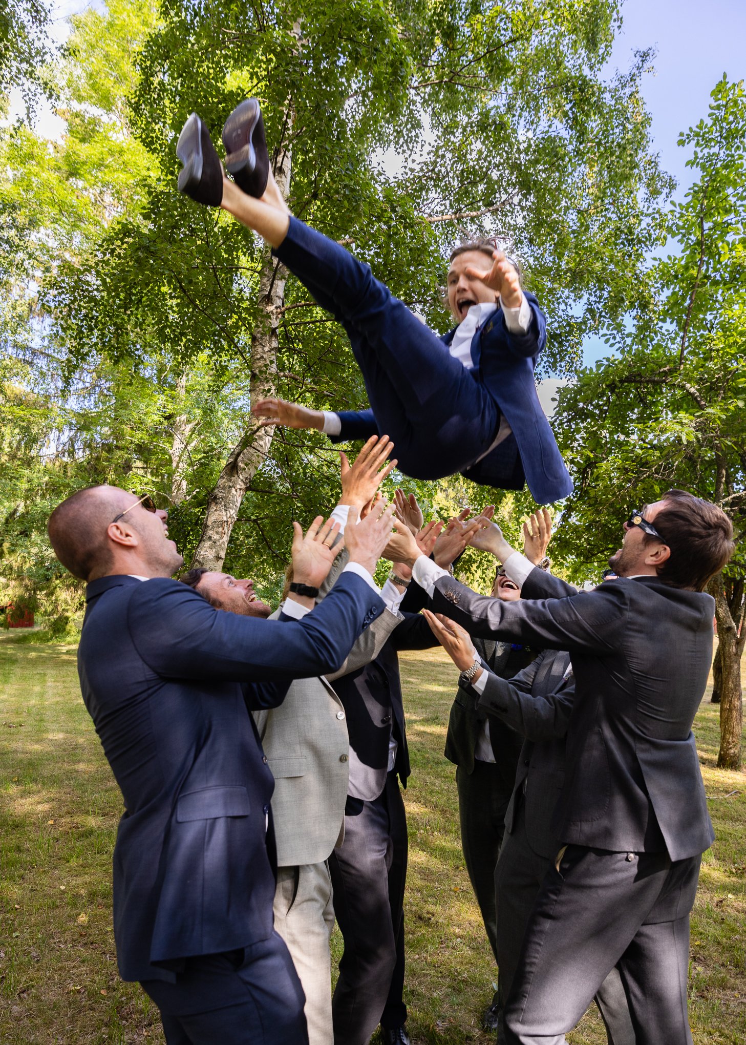 Wedding guests tossing a smiling groom into the air outdoors.