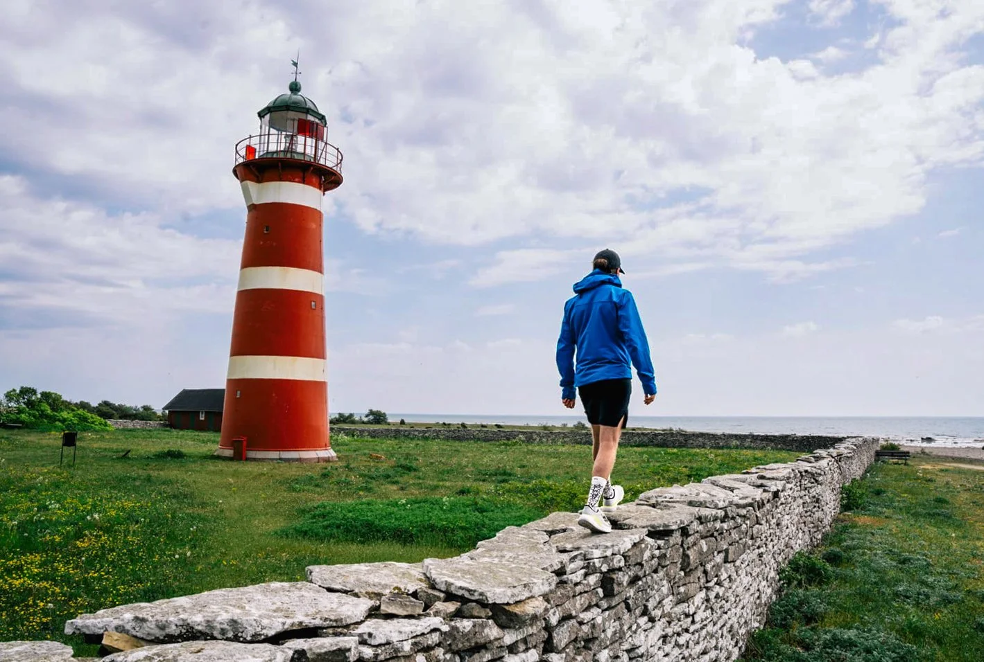 A person walking along a stone wall beside a red-and-white lighthouse.
