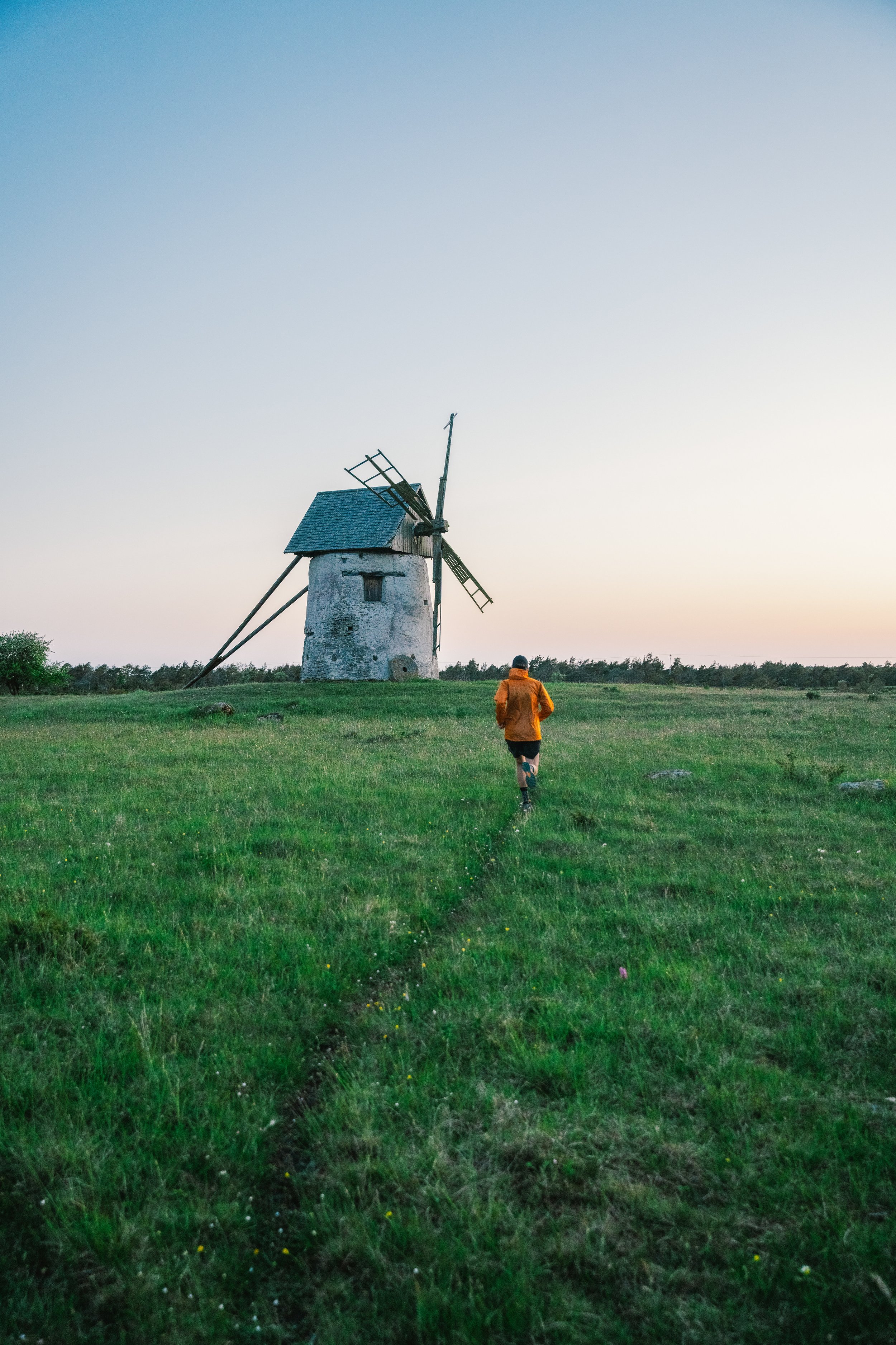 A runner in an orange jacket approaching an old windmill across a grassy field.