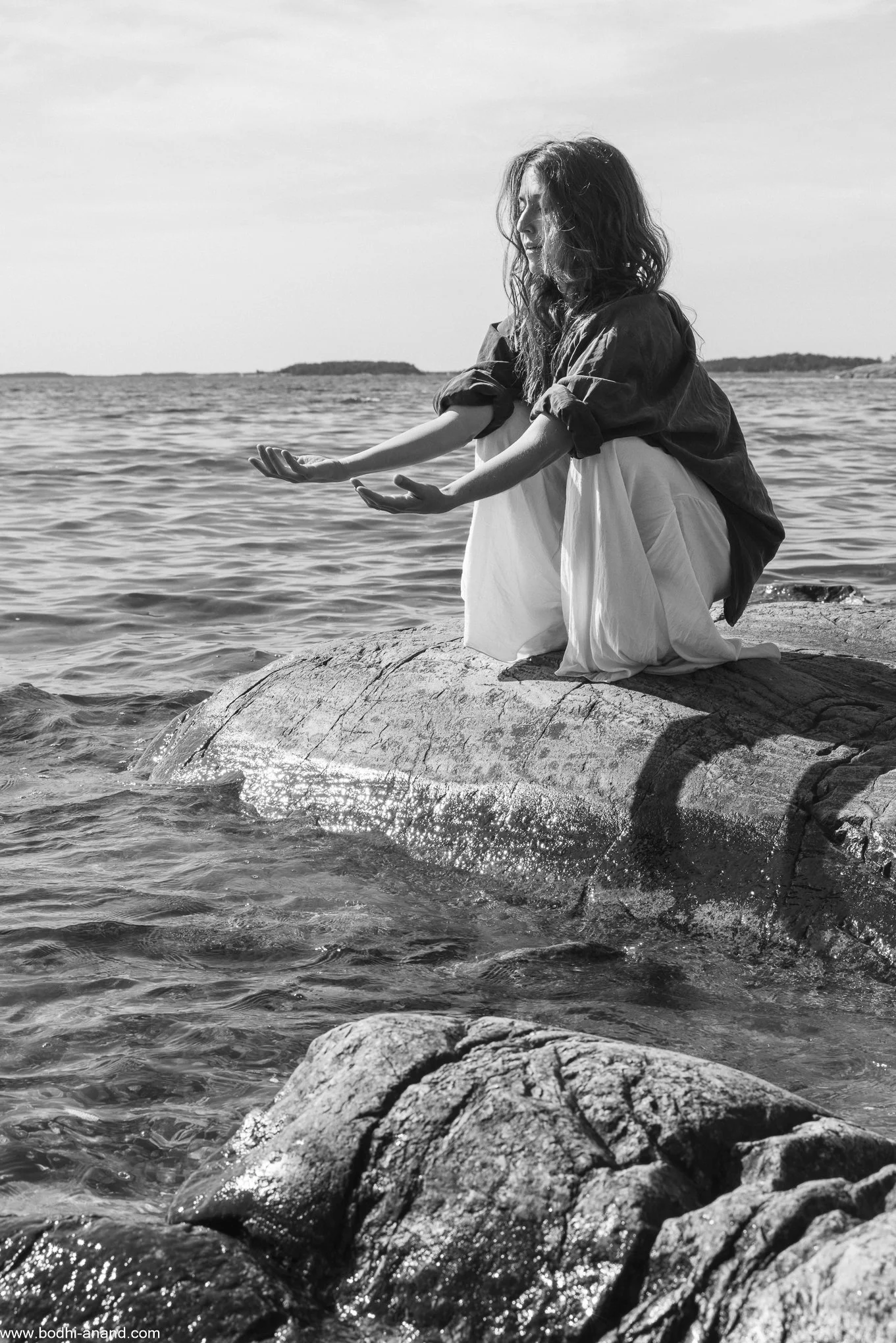 Black-and-white portrait of a woman sitting on rocks beside the sea.