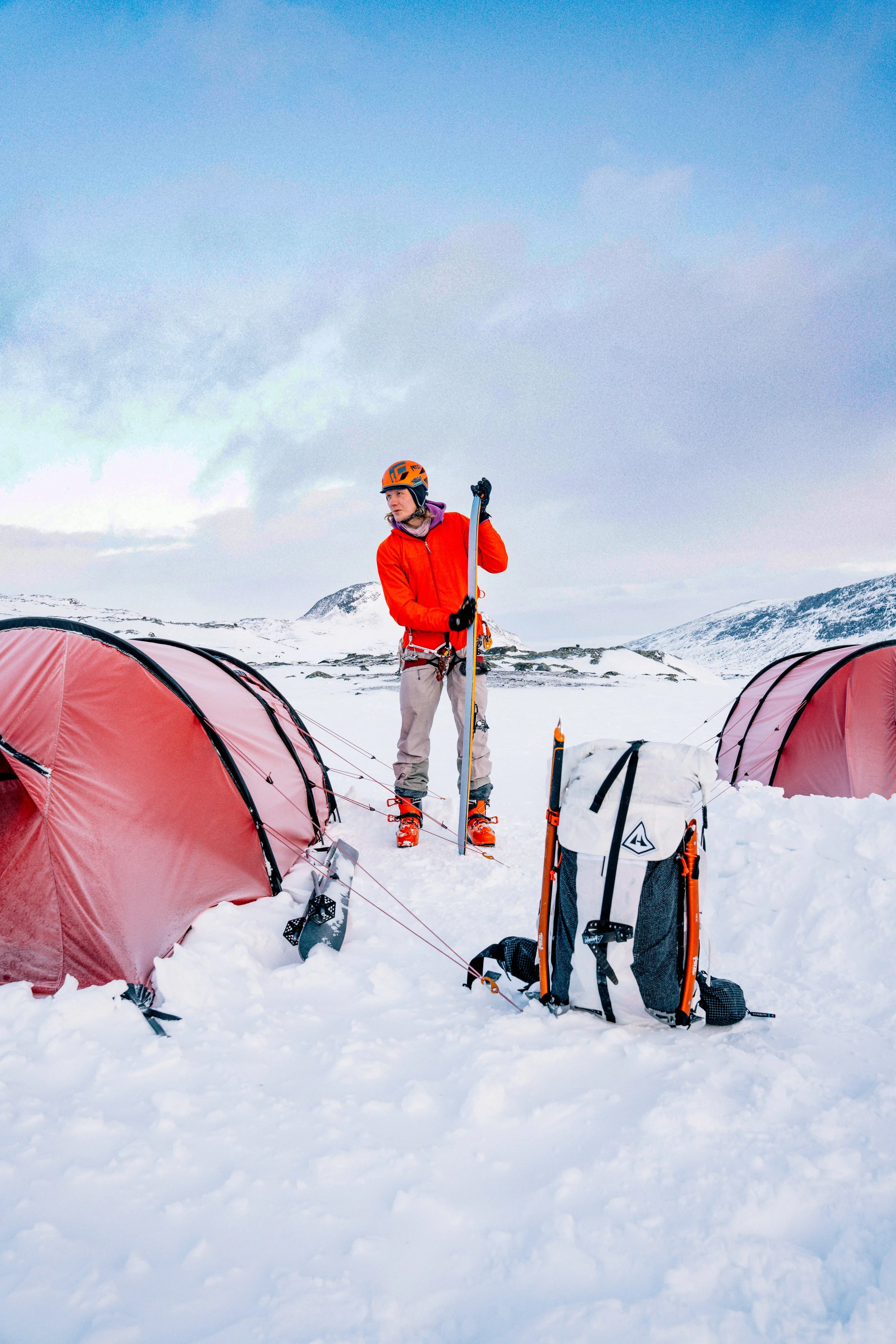 A skier standing between tents and equipment at a snowy mountain campsite.