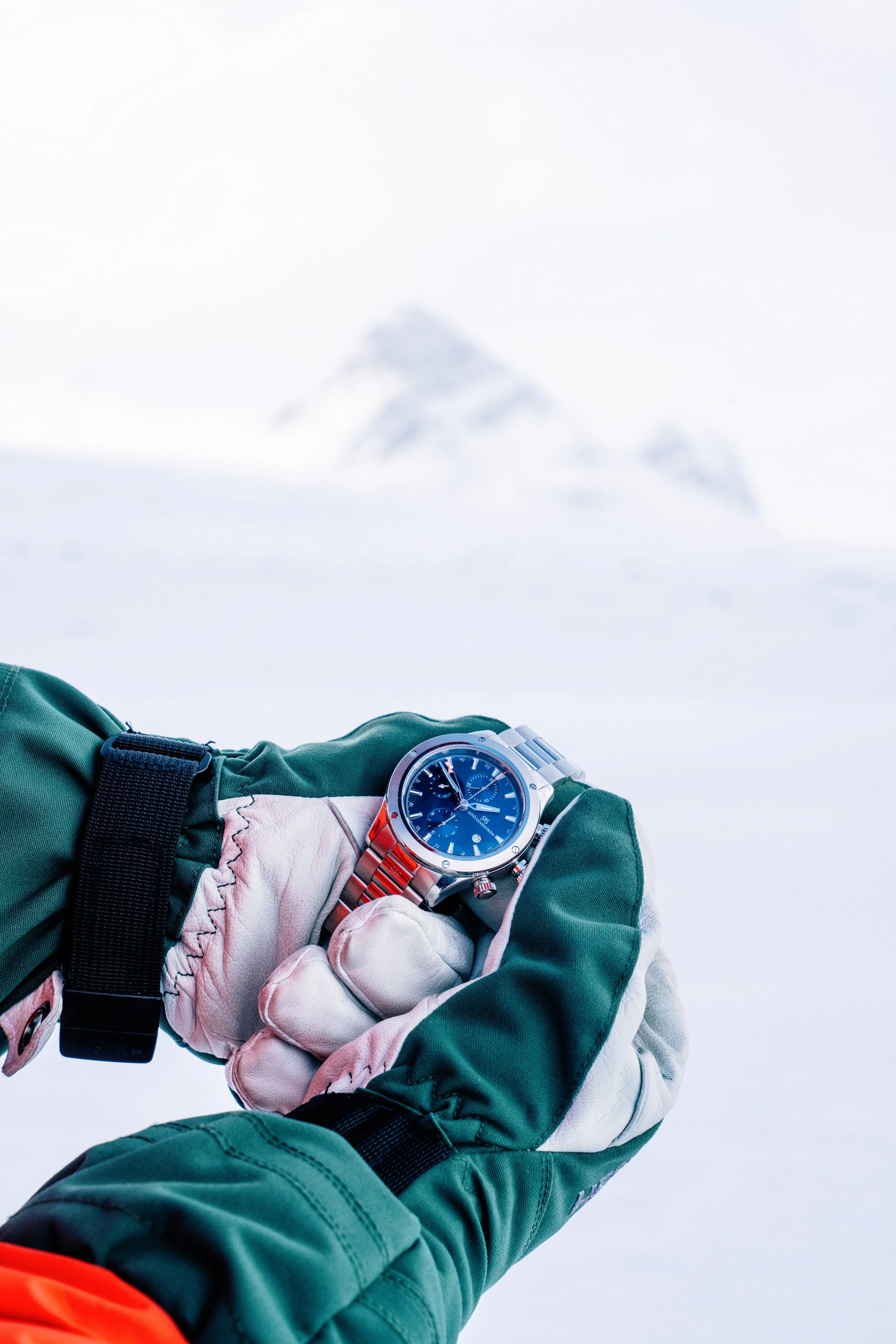 A blue-dial wristwatch resting on a gloved hand in snowy mountains.