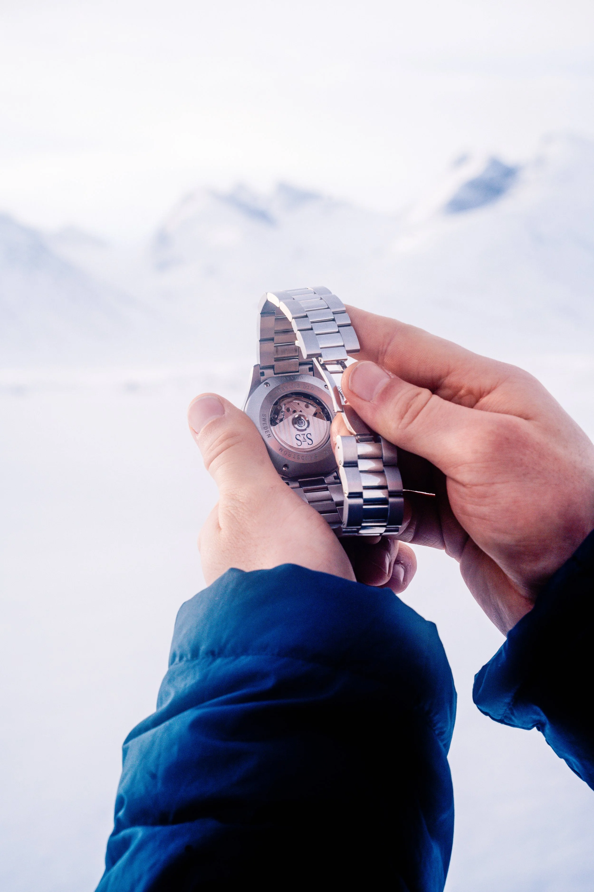 Hands holding a metal-bracelet wristwatch against a snowy mountain landscape.