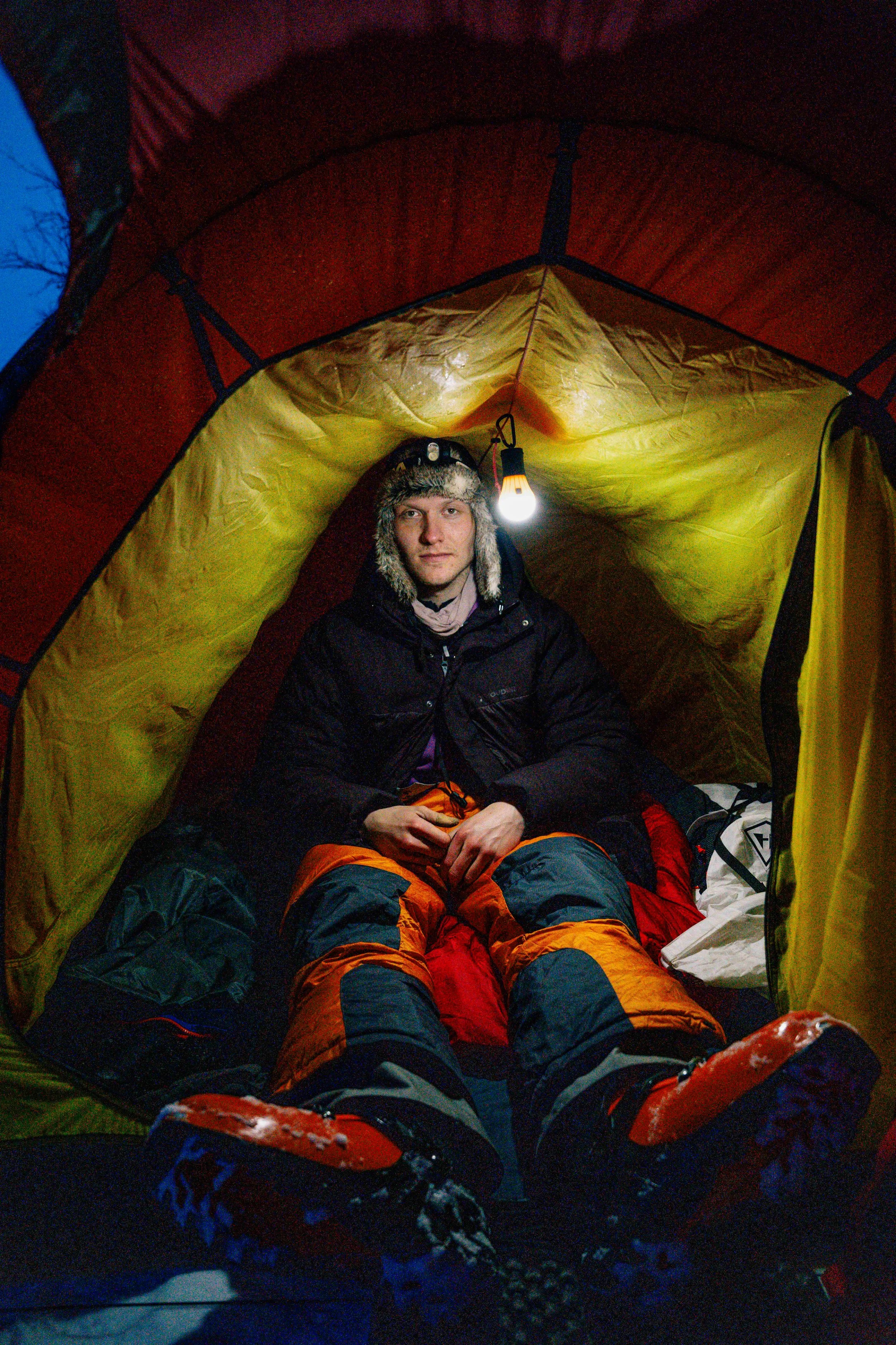 A person in winter clothing sitting inside a lit tent.