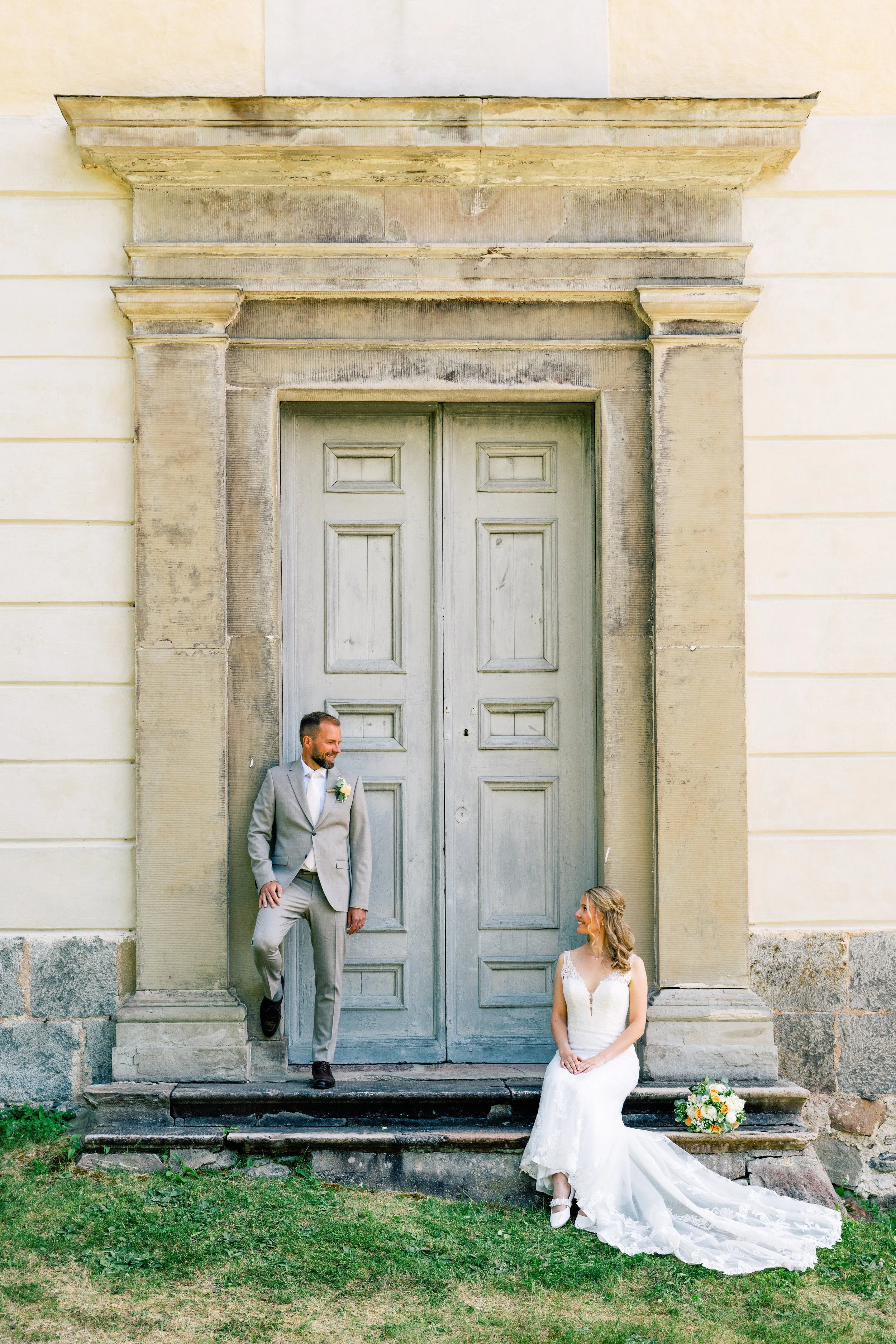 Bride and groom outside a tall doorway, with the bride seated on the steps.