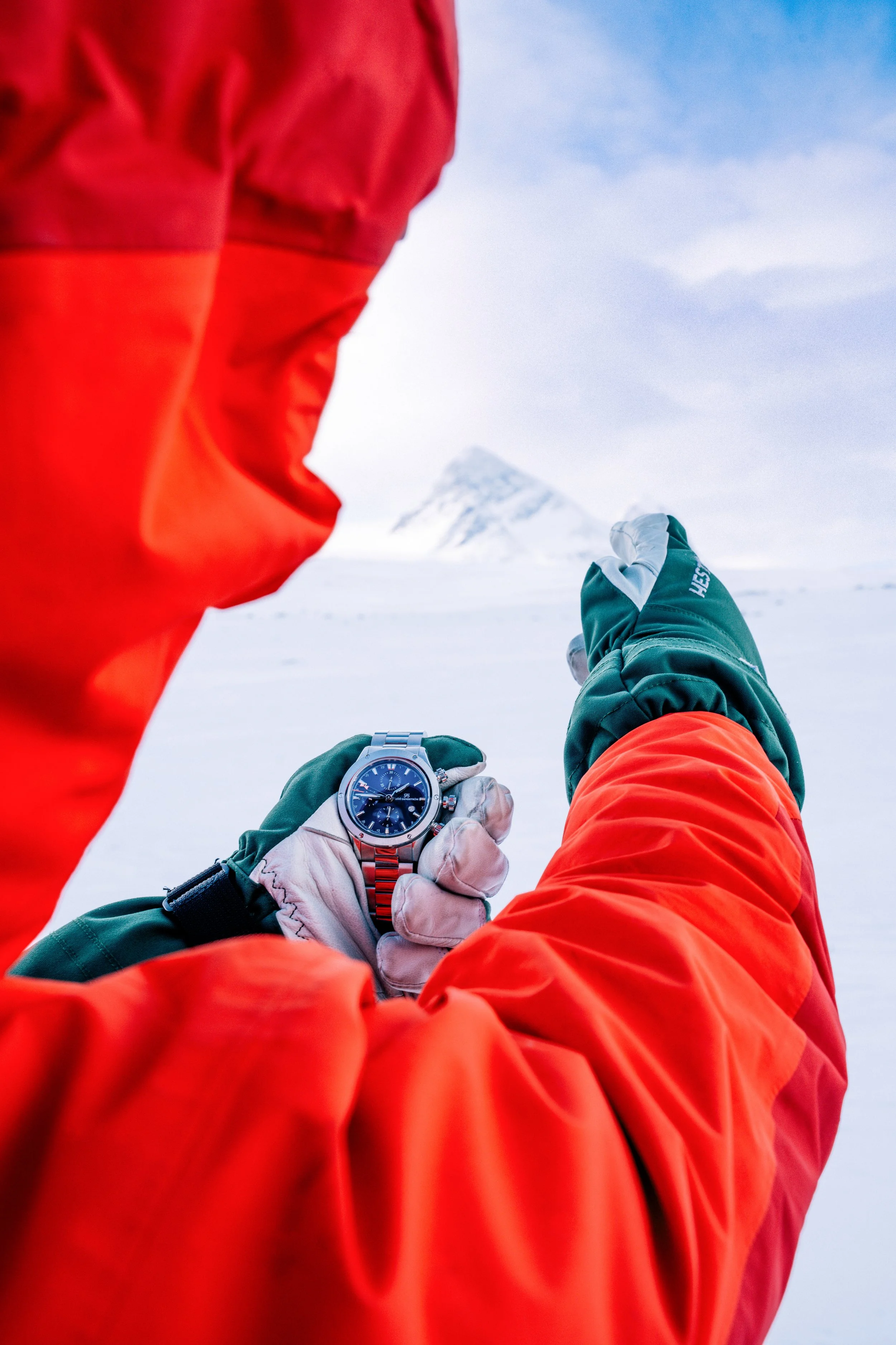 A person in an orange jacket comparing a wristwatch with the mountain view.