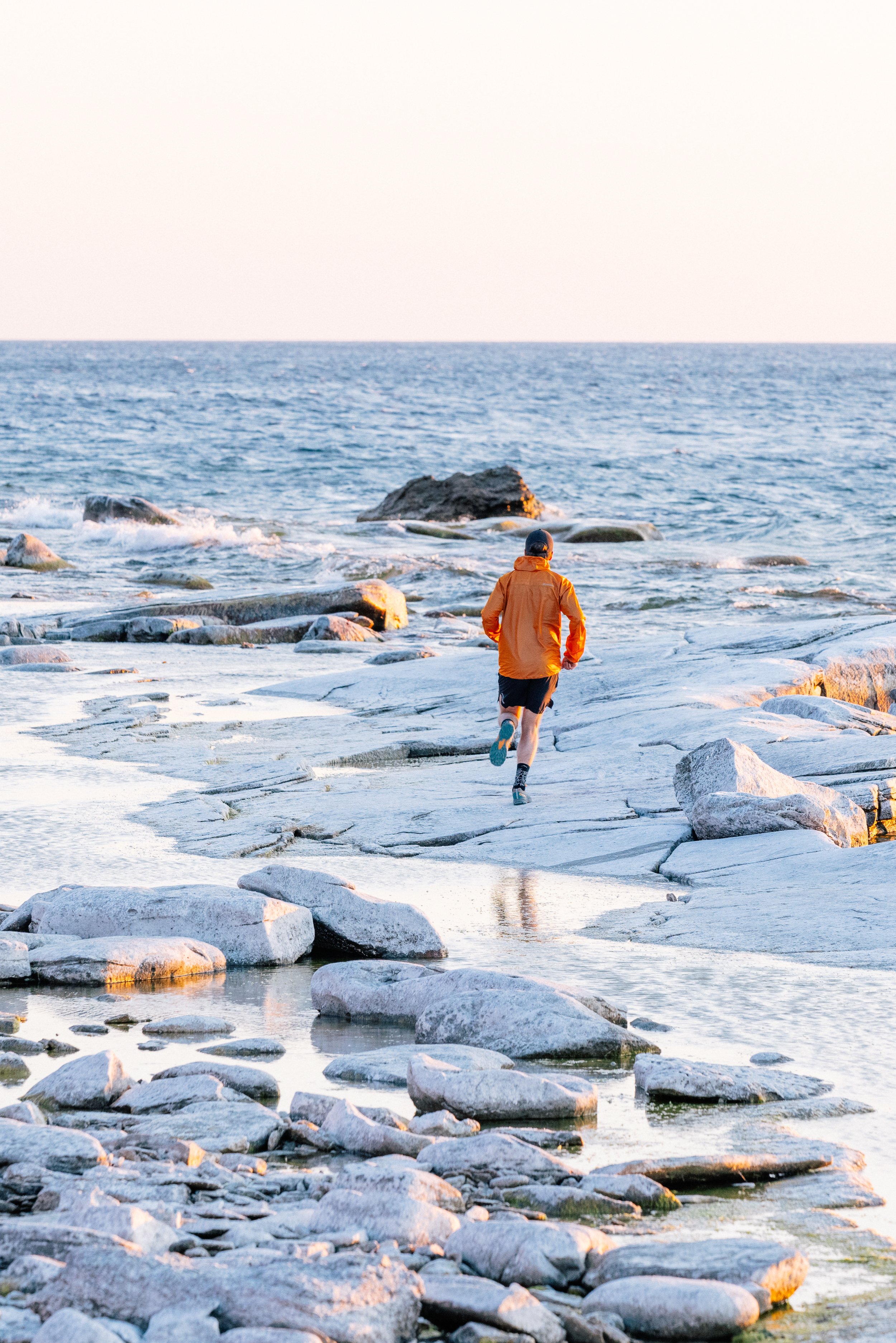 A runner in an orange jacket crossing rocks beside the sea.