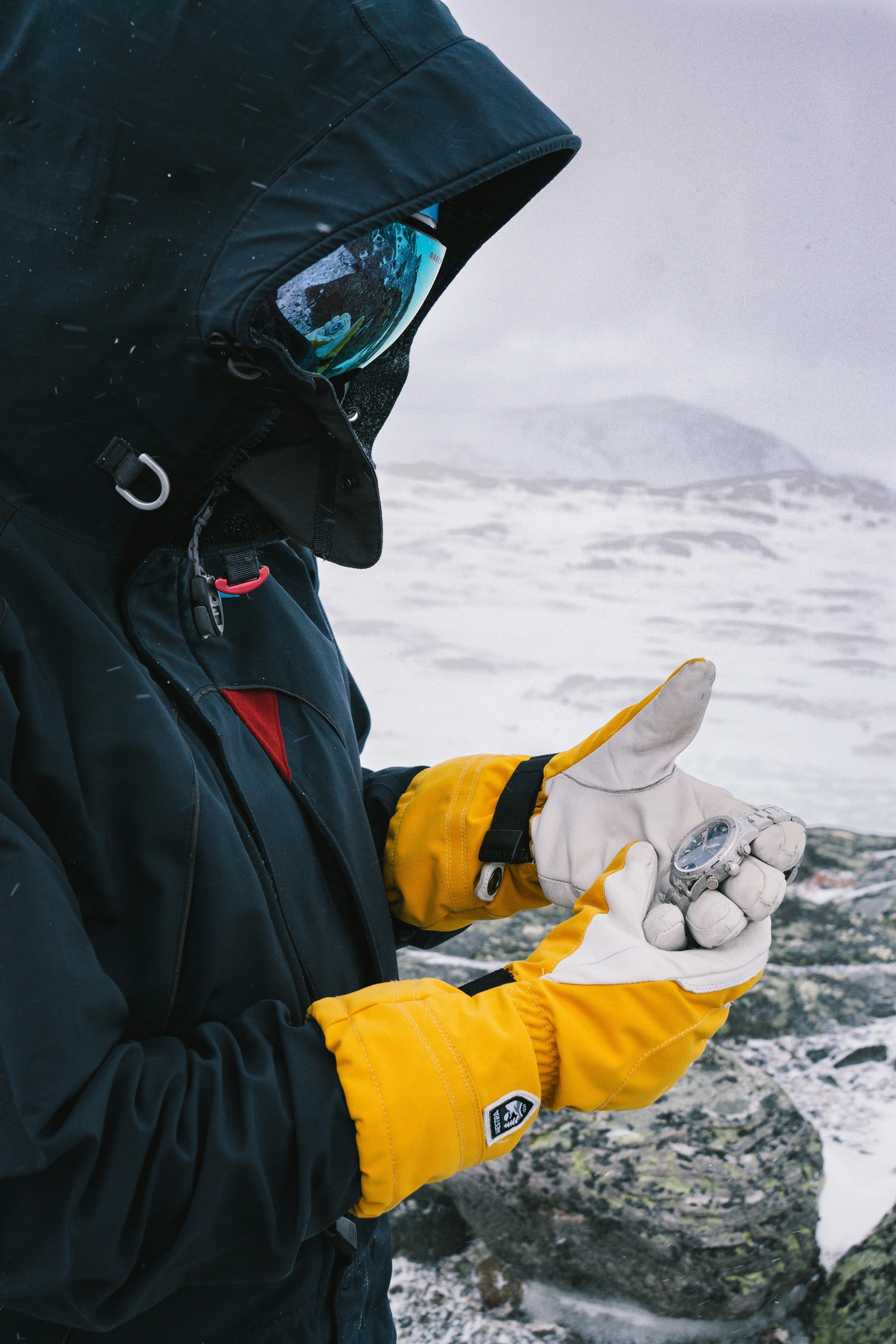 A hooded person inspecting a wristwatch with gloved hands in the mountains.