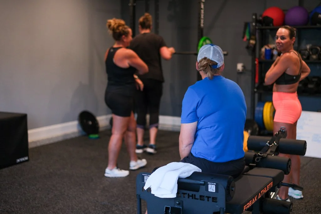 Four women and a trainer in a gym. One woman sits on a workout bench, while the others stand and chat. The gym, in saint augustine, fl has various fitness equipment, including medicine balls and weights.