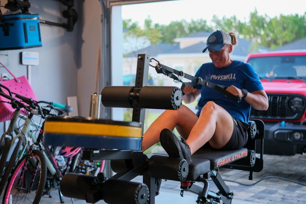 A woman smiling and exercising on a rowing machine in a saint augustine, fl garage gym, with bicycles and a red vehicle in the background.