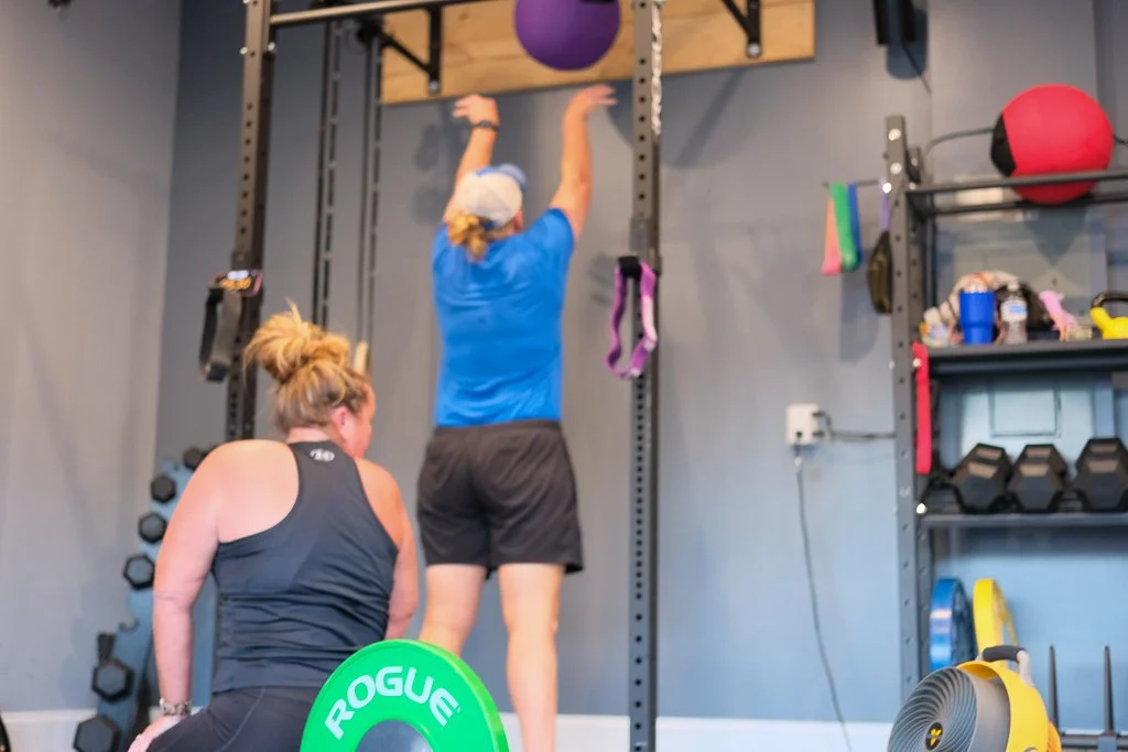 2 women working out in a gym, with one woman kneeling and the other performing wall balls.