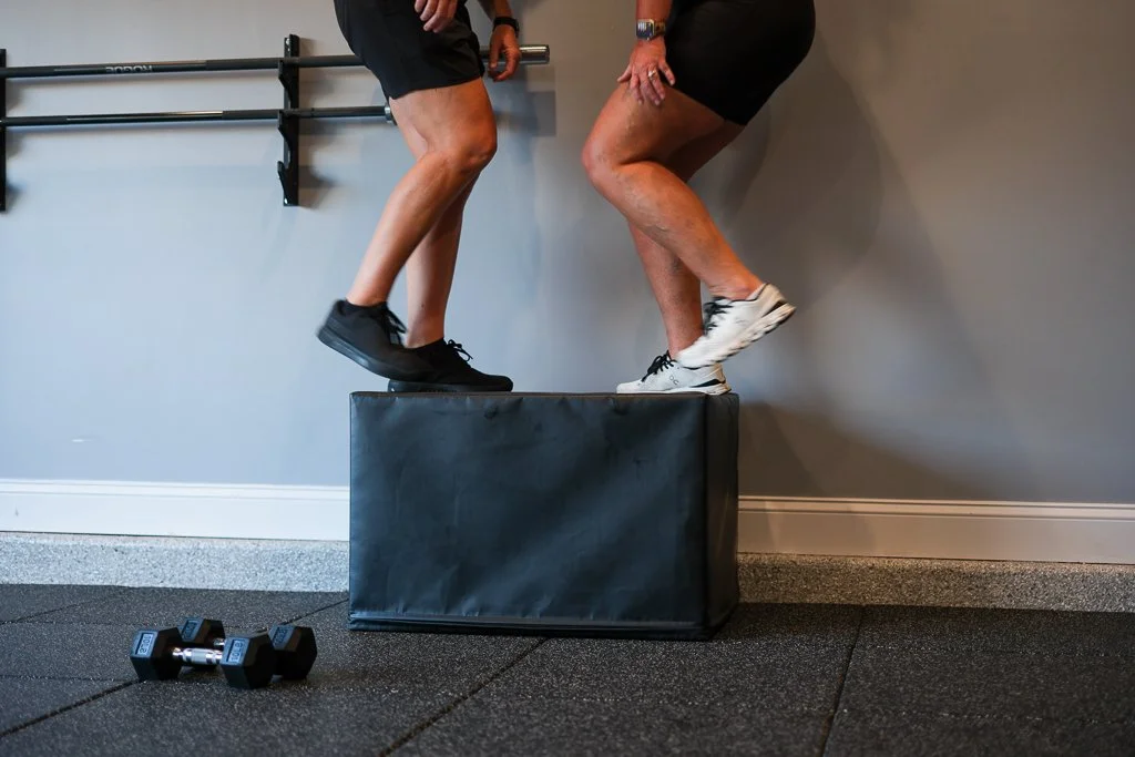 Two people balancing on a black padded box in a gym, with dumbbells and a wall-mounted barbell in the background.
