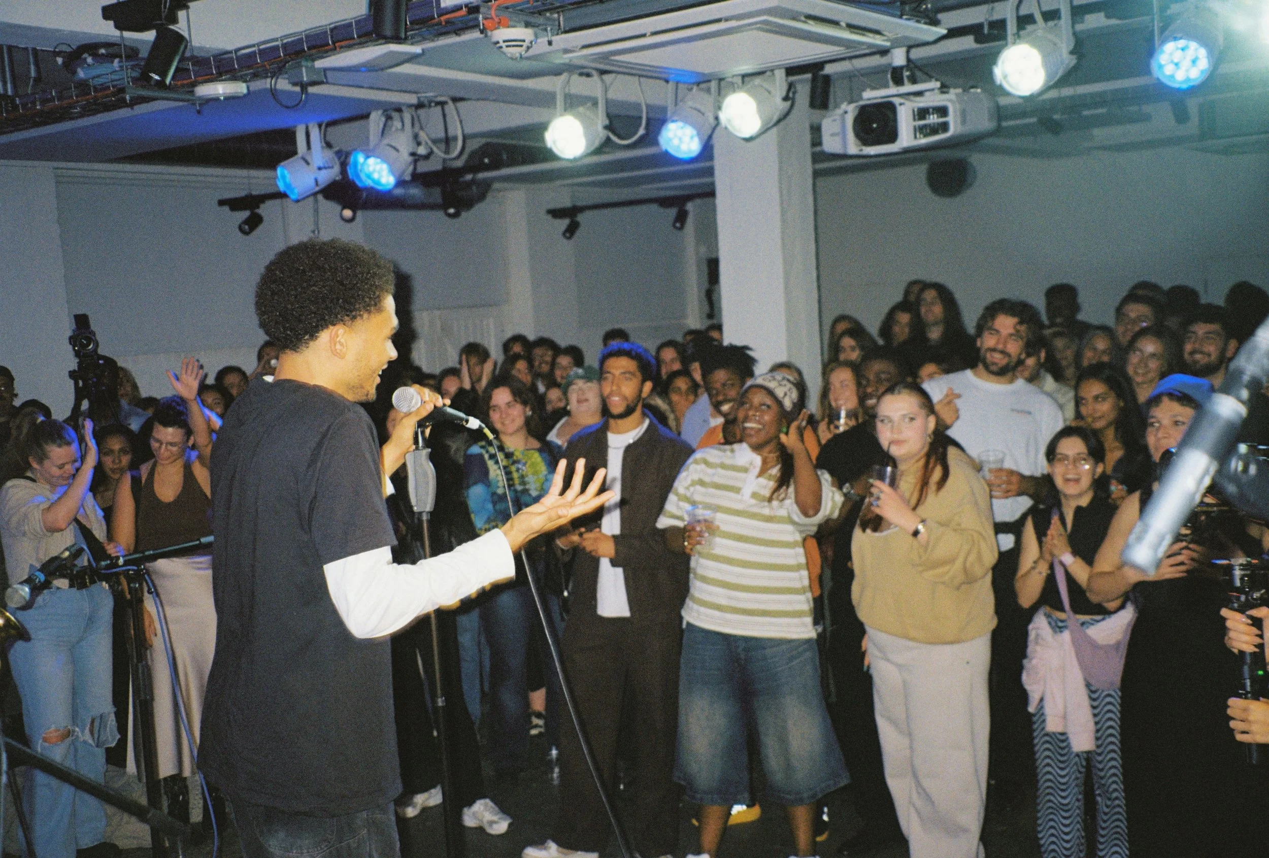 A young man with curly hair speaking into a microphone at a crowded indoor event, with an audience clapping and smiling.