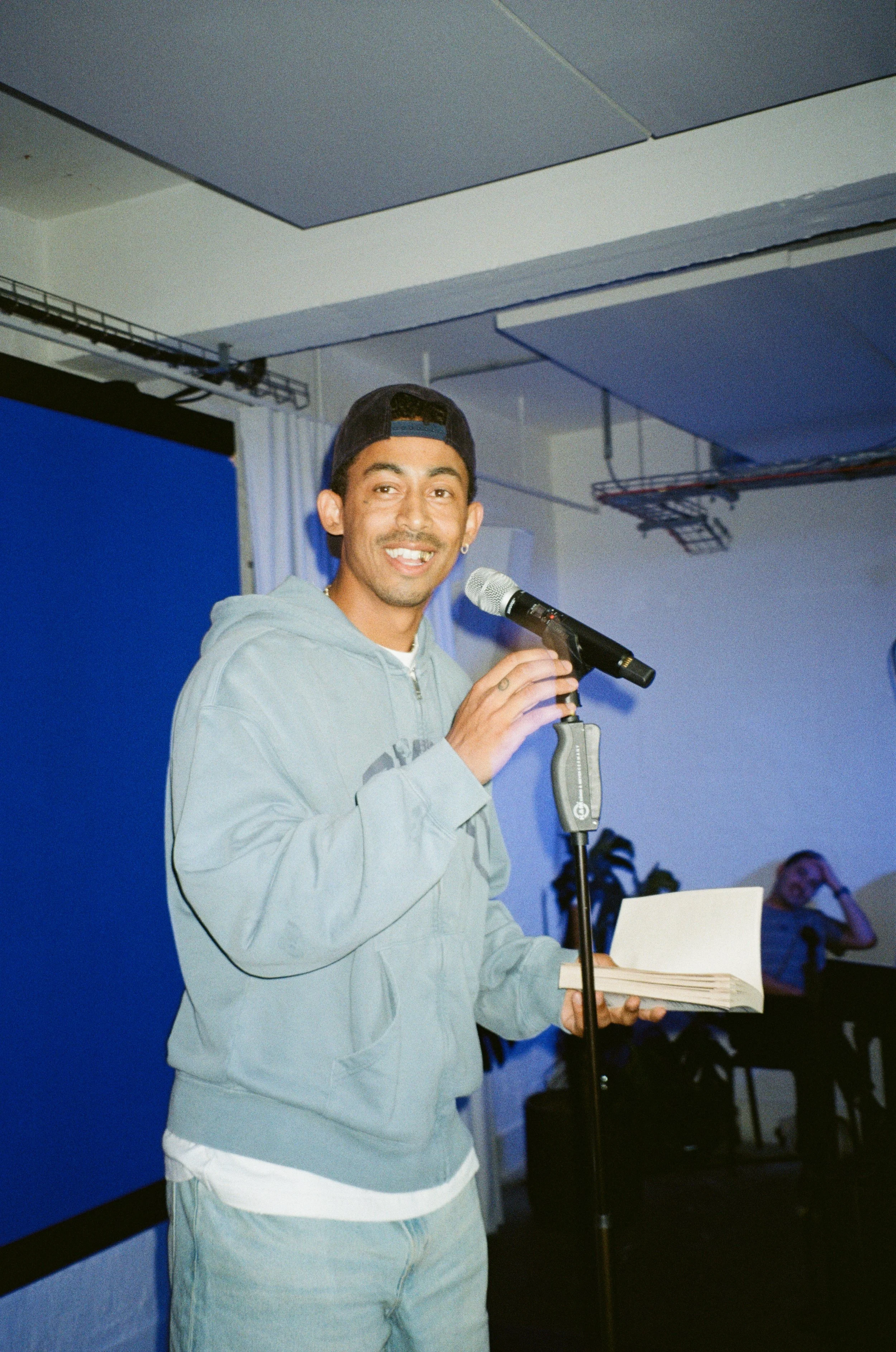 A young man with a gray hoodie, holding papers, speaking into a microphone on a stand in front of a blue screen in a room with a woman sitting at a table in the background.