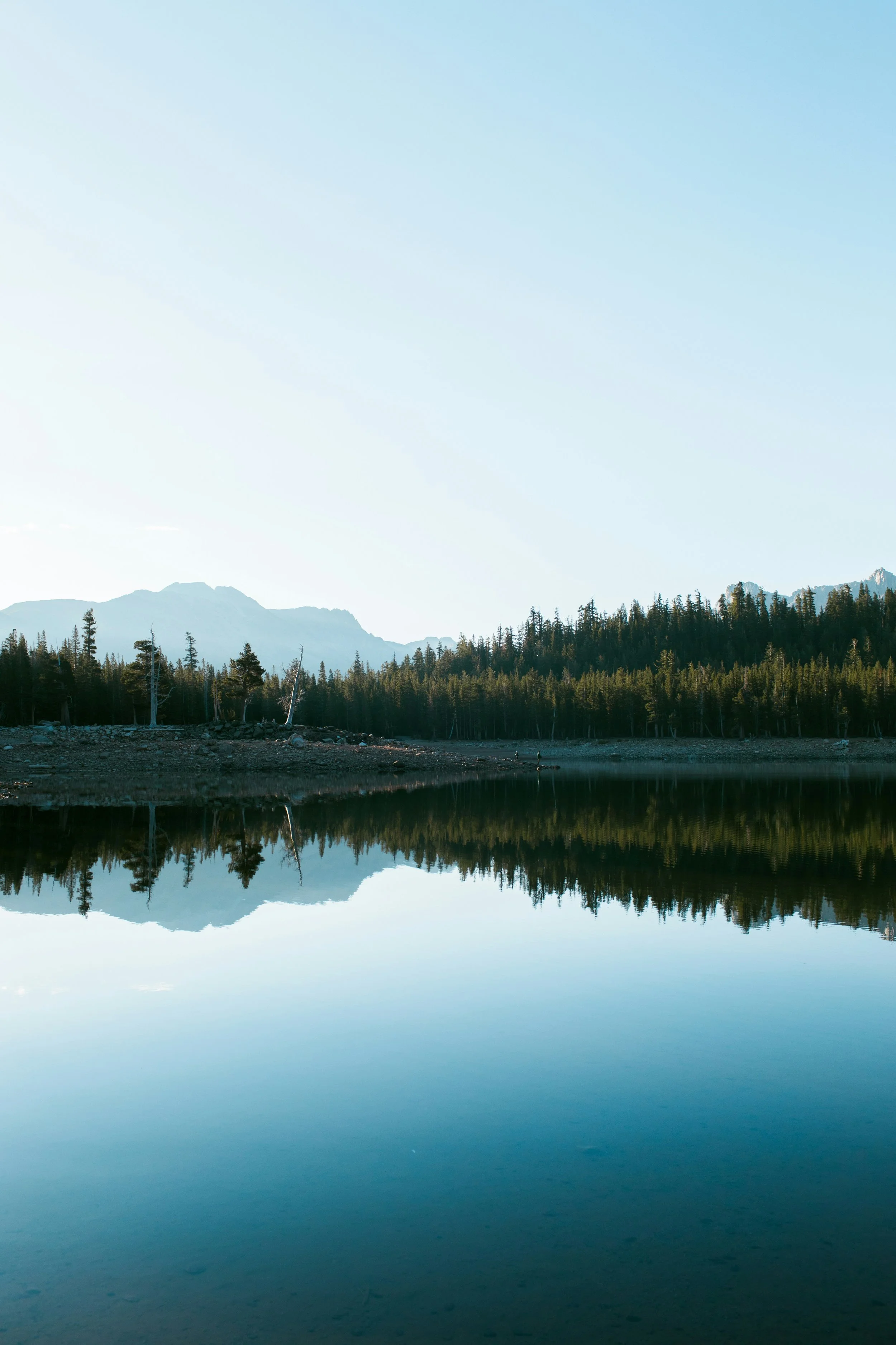 A calm mountain lake with a mirror-like reflection of trees and mountains in the water under a clear, light blue sky.