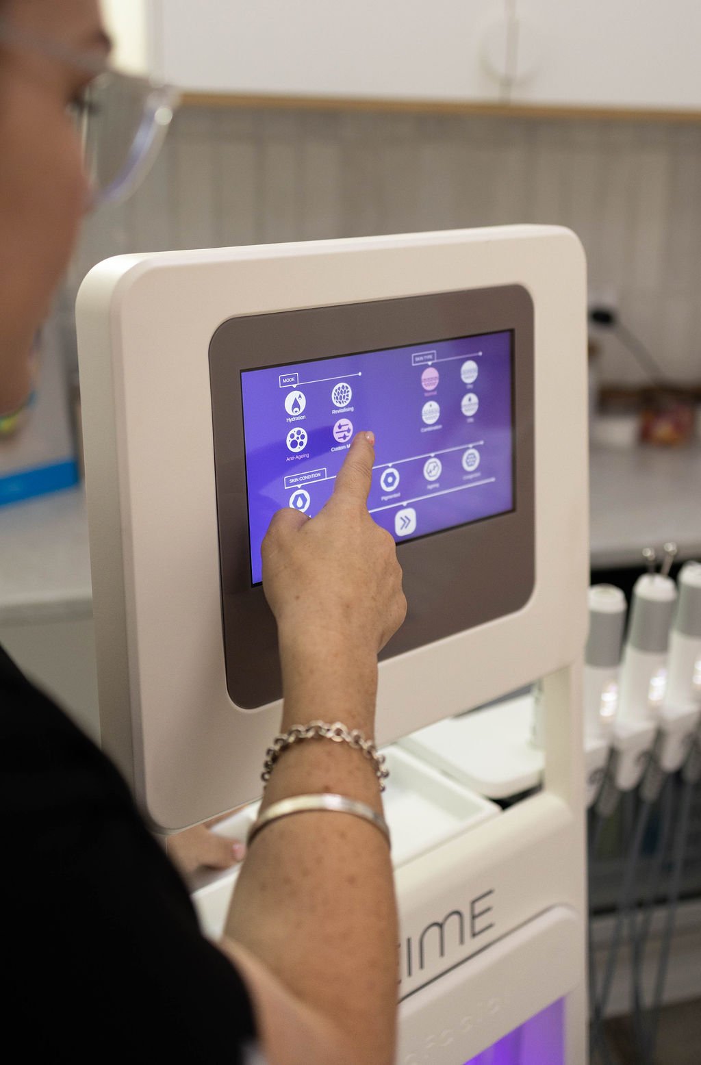 A person using a skincare or cosmetic treatment kiosk with a touchscreen interface, selecting options related to hydration, anti-aging, and skin condition.