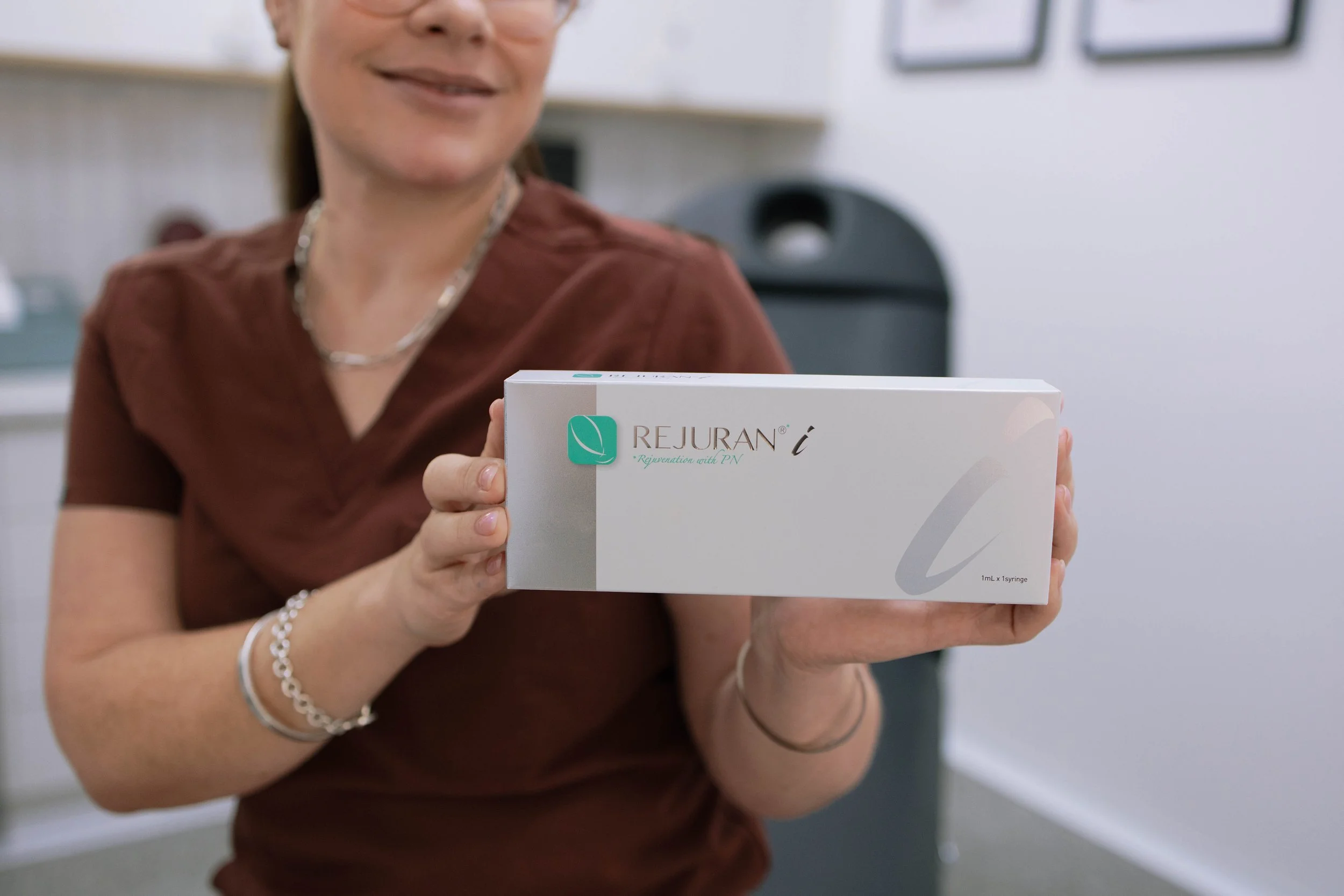 A woman in a brown shirt holding a box of Rejuran skincare product, smiling.