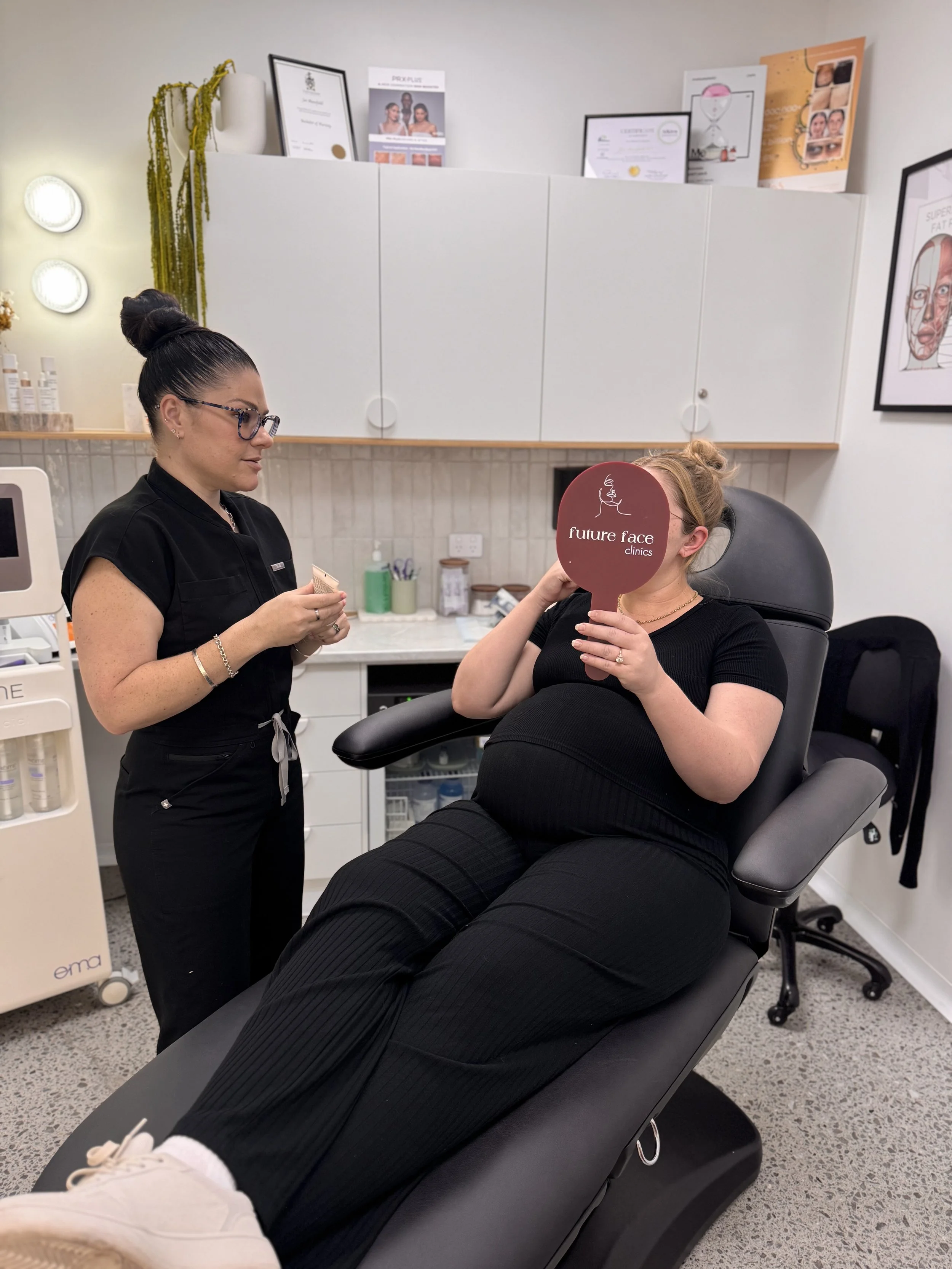 A woman sitting in a medical chair, holding a mirror with the words 'future face clinics' on it, in a clinic consultation room. A medical professional is standing nearby, talking to her. The room has medical equipment, certificates on the wall, and decorative plants.