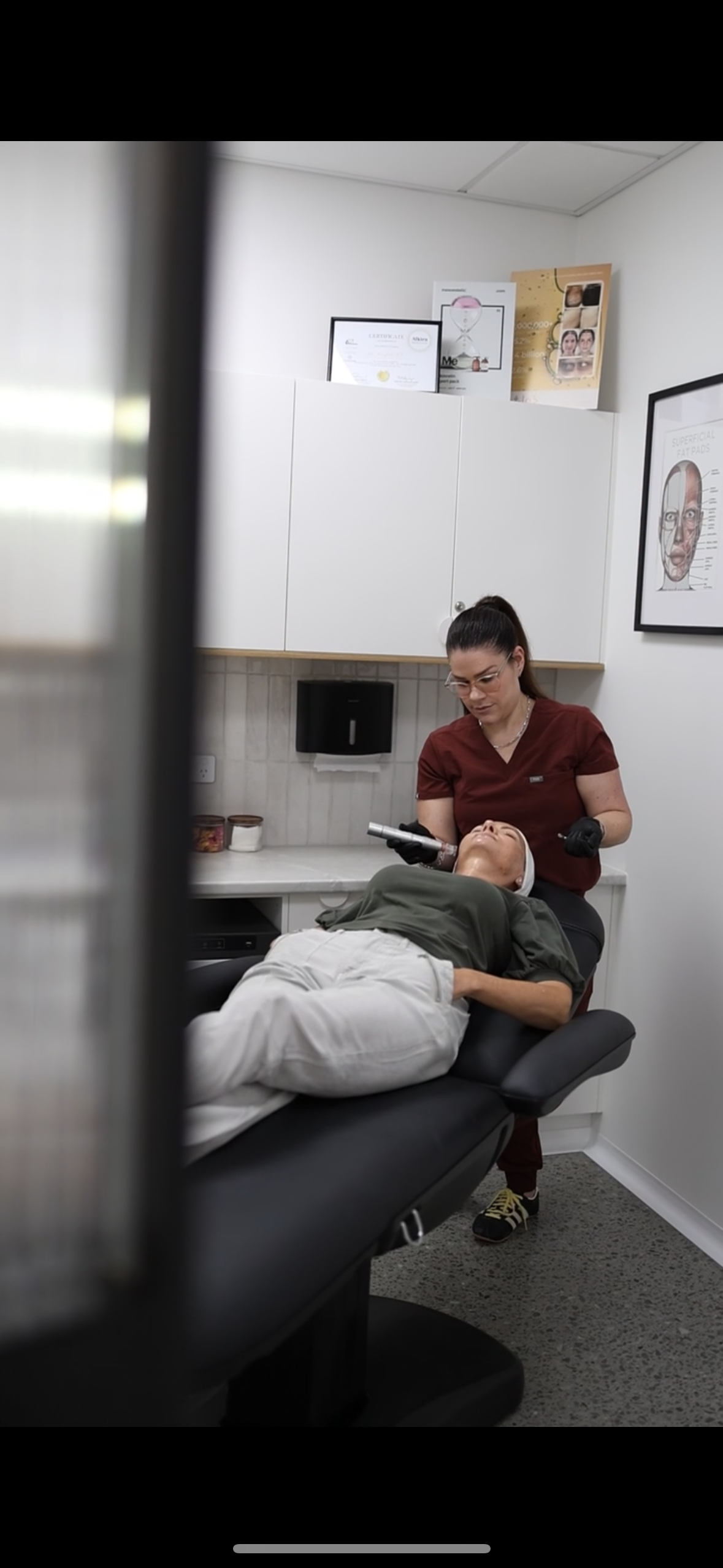 A woman receiving a facial treatment from a skincare professional in a clinic room with white cabinets and framed certificates.