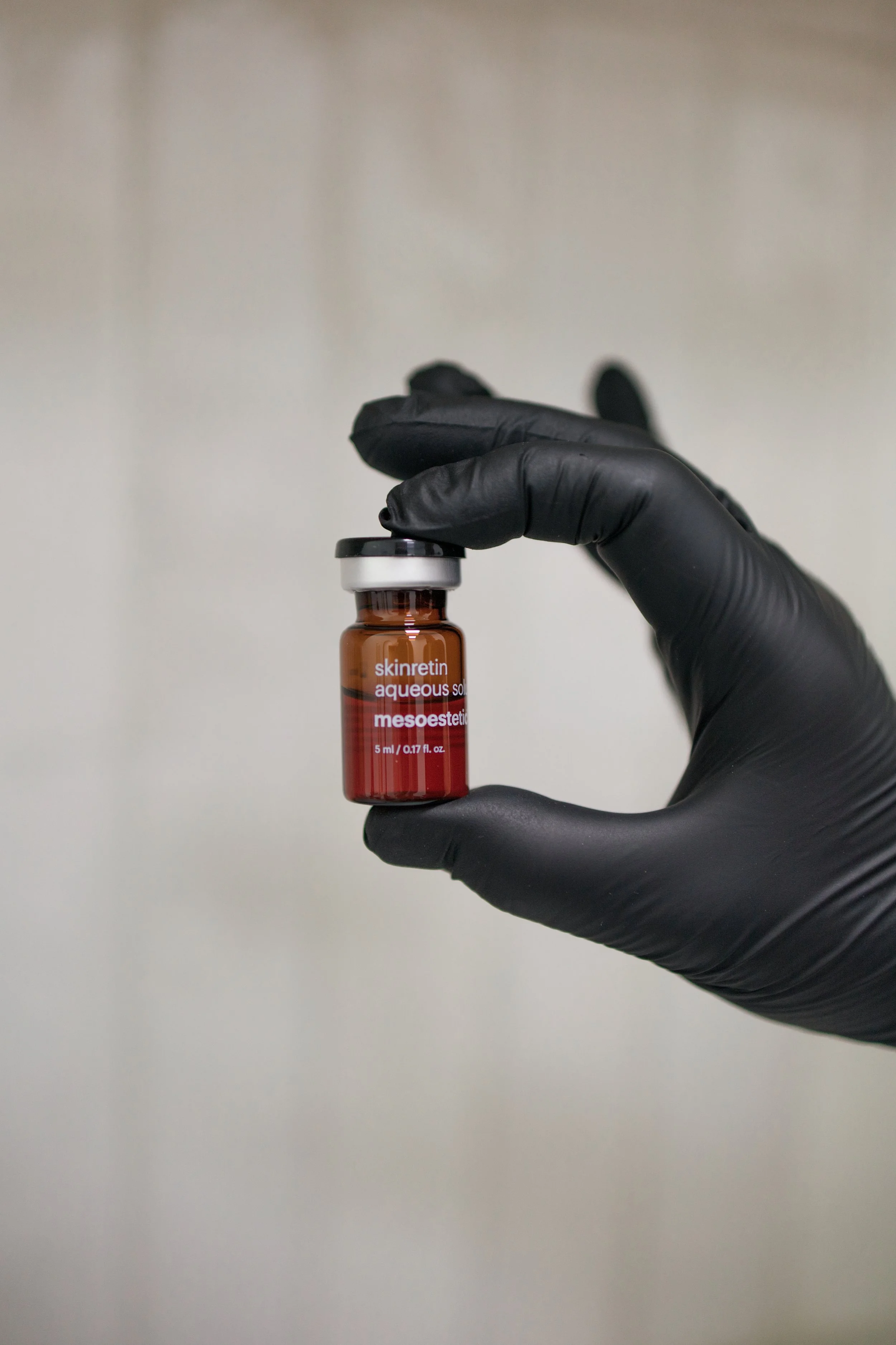 A hand wearing a black glove holding a small vial labeled 'skinre tin aqueous sol mesoestetic' against a neutral background.
