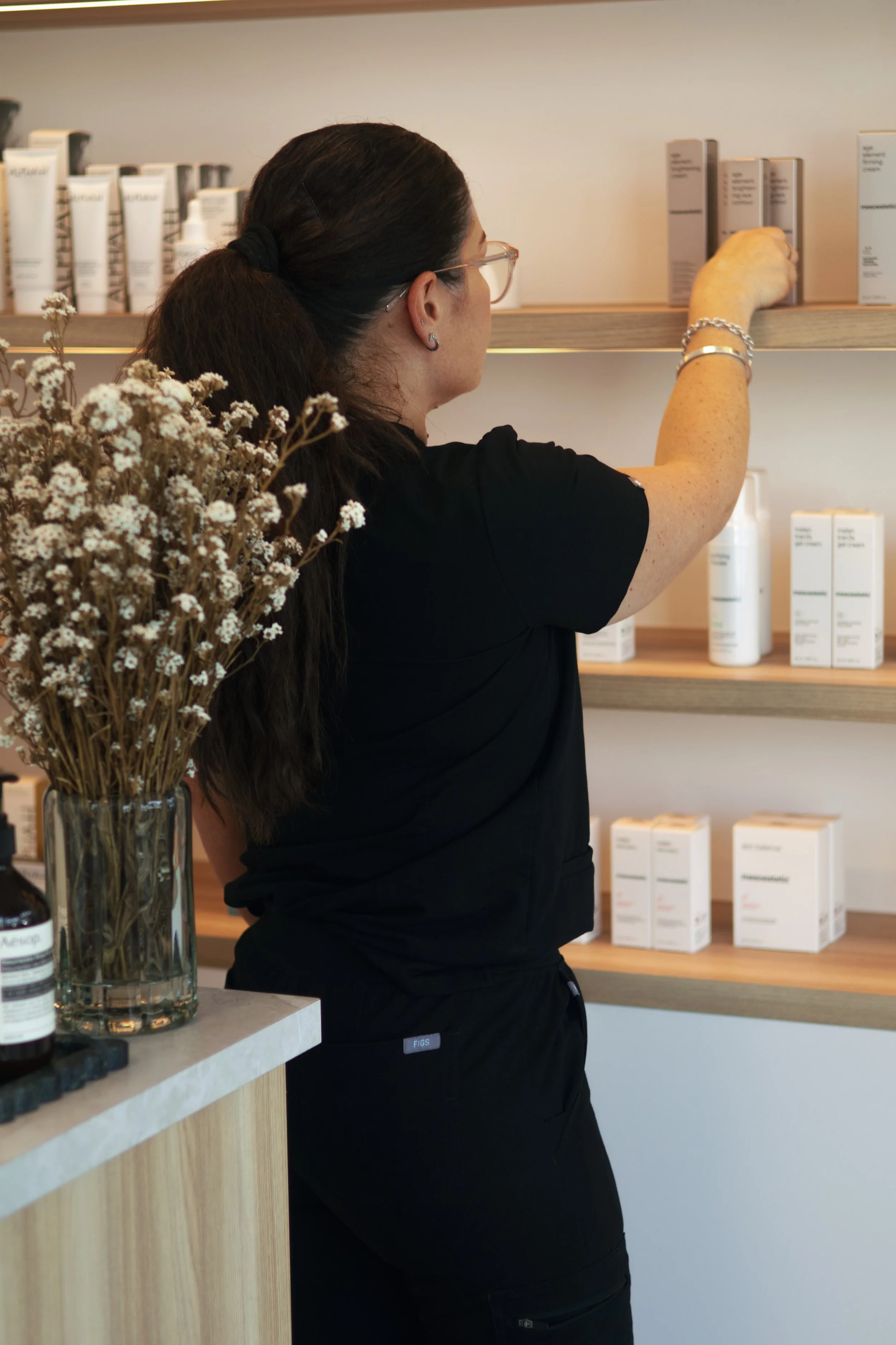 A woman with dark hair in a ponytail, wearing glasses and a black shirt, is reaching for products on a wooden shelf in a store. There are white and gray boxes and bottles on the shelves. A glass vase with dried white flowers is on a counter nearby.